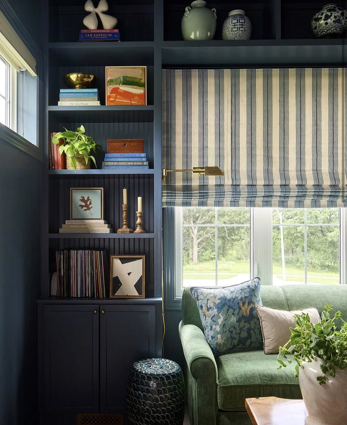 A living room corner with a dark blue bookshelf filled with books, decorative items, and framed artwork. A green sofa with patterned throw pillows in front of a window with striped blue and yellow Roman blinds, overlooking a green outdoor area.
