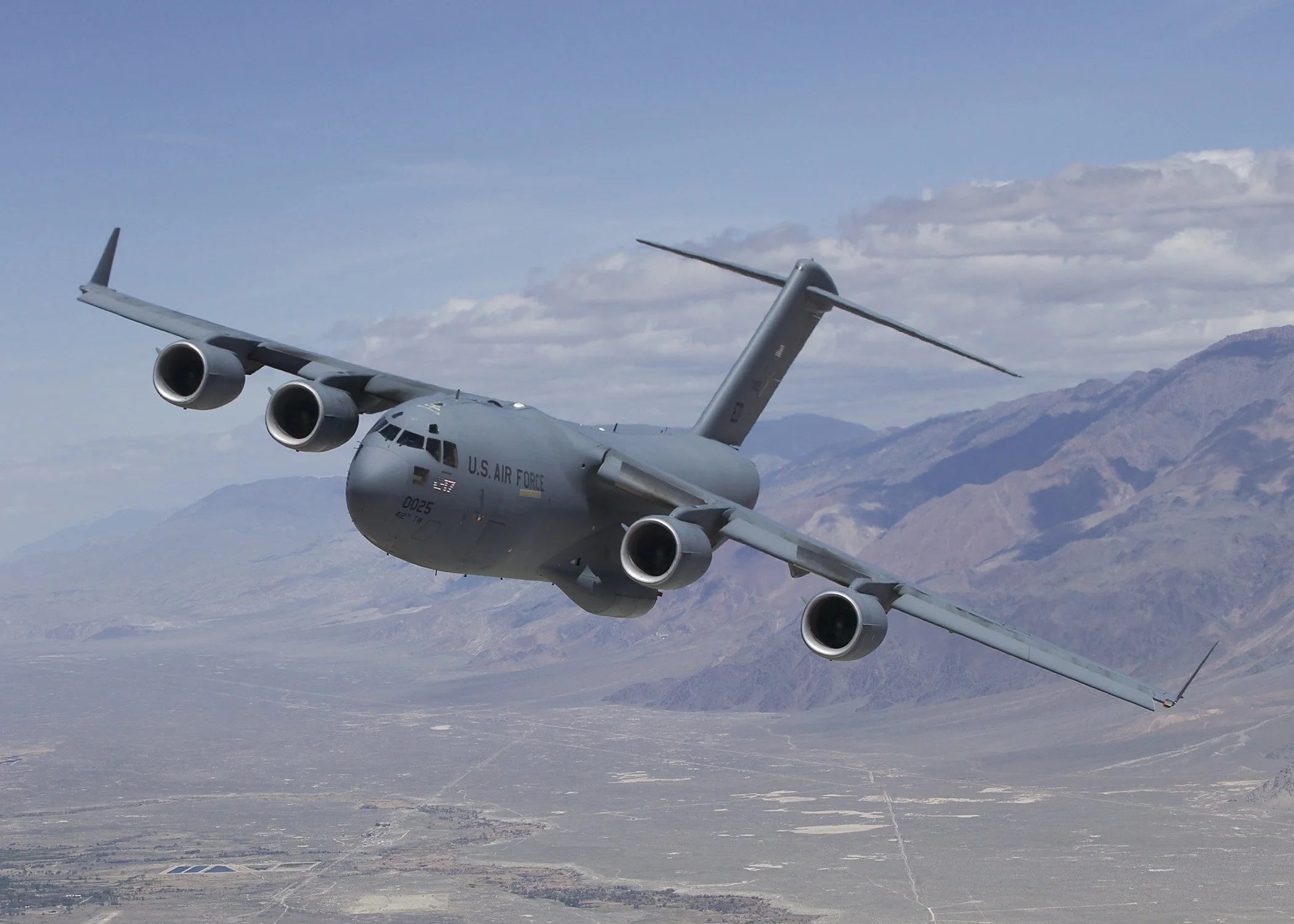 A U.S. Air Force cargo plane flying over a desert landscape with mountains in the background.