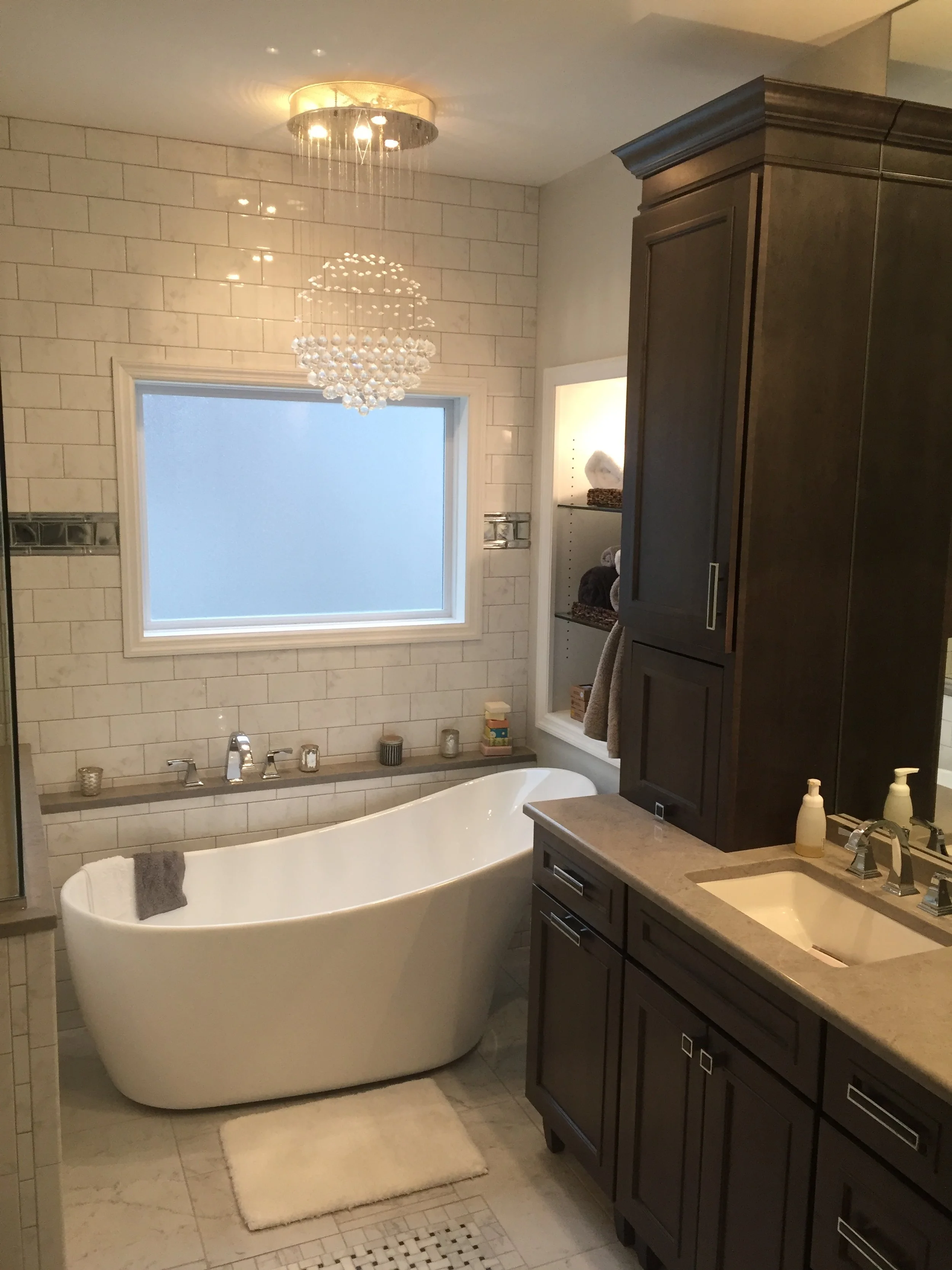Modern bathroom with a white freestanding bathtub, dark wooden cabinets, a large window with frosted glass, and a chandelier hanging from the ceiling.