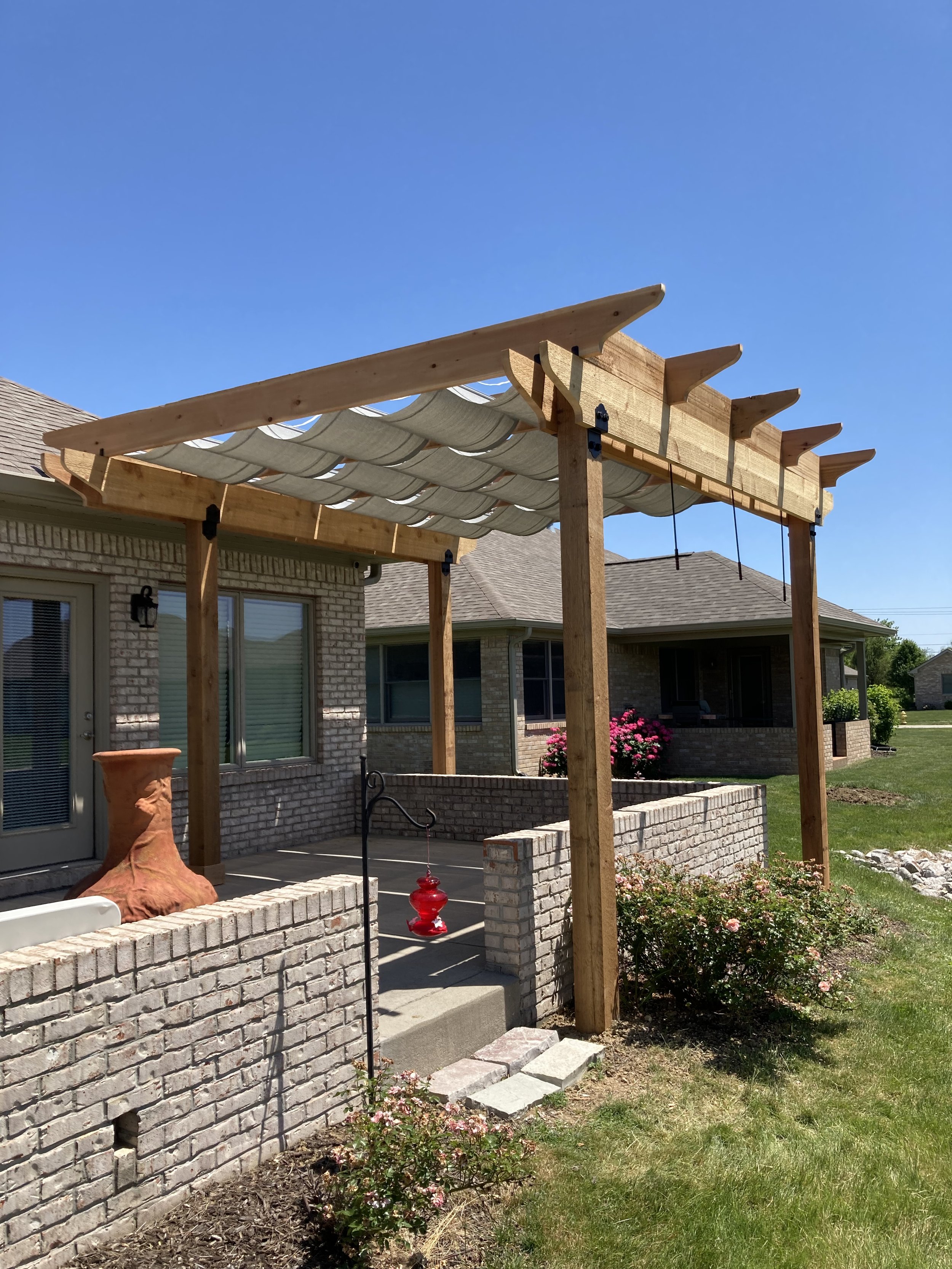 Backyard patio with a wooden pergola under construction, brick wall, potted plants, and a clear blue sky.