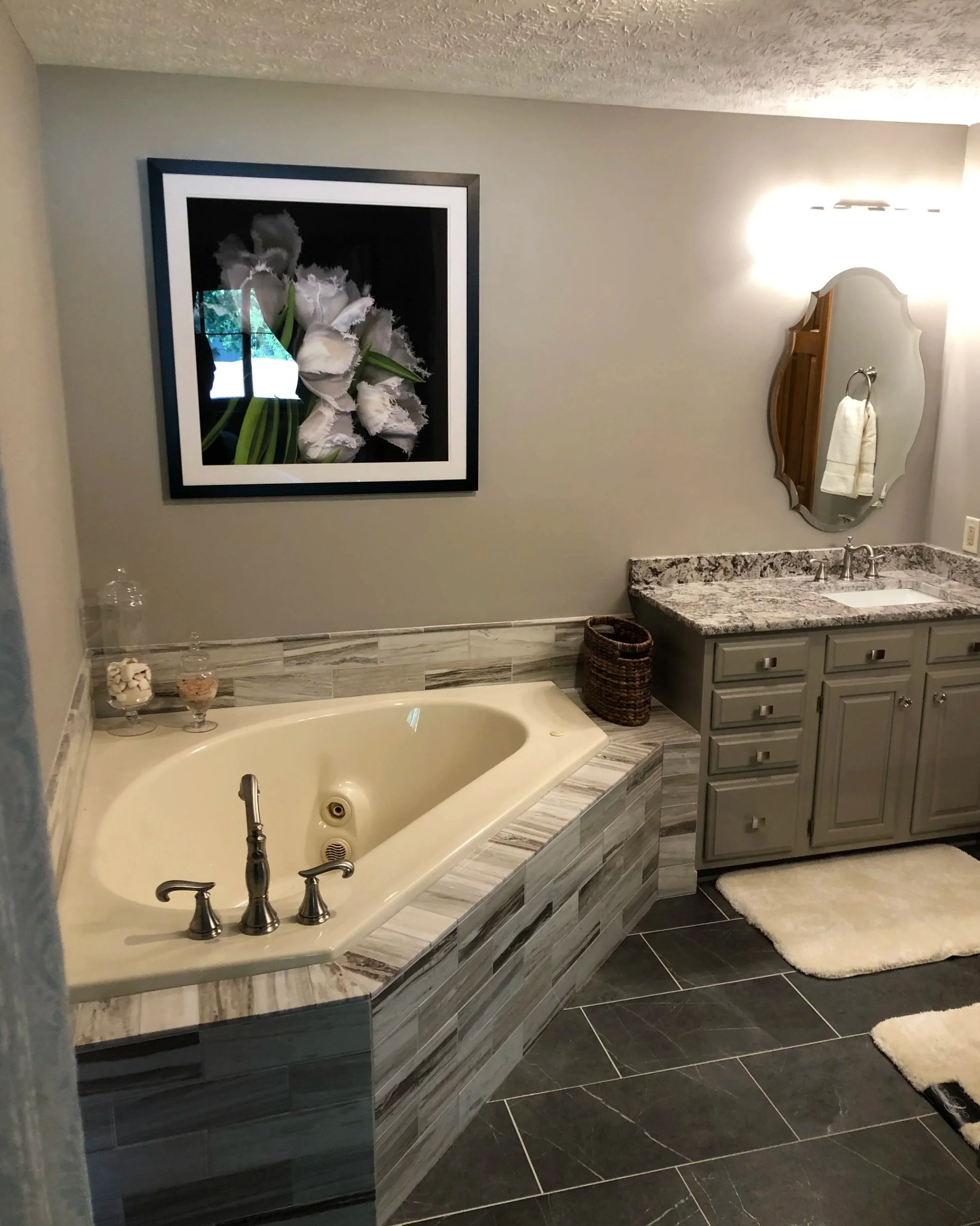 A bathroom with a corner bathtub surrounded by gray and beige tiles, a gray vanity with a granite countertop, a decorative mirror, and a large framed picture of white flowers on the wall.