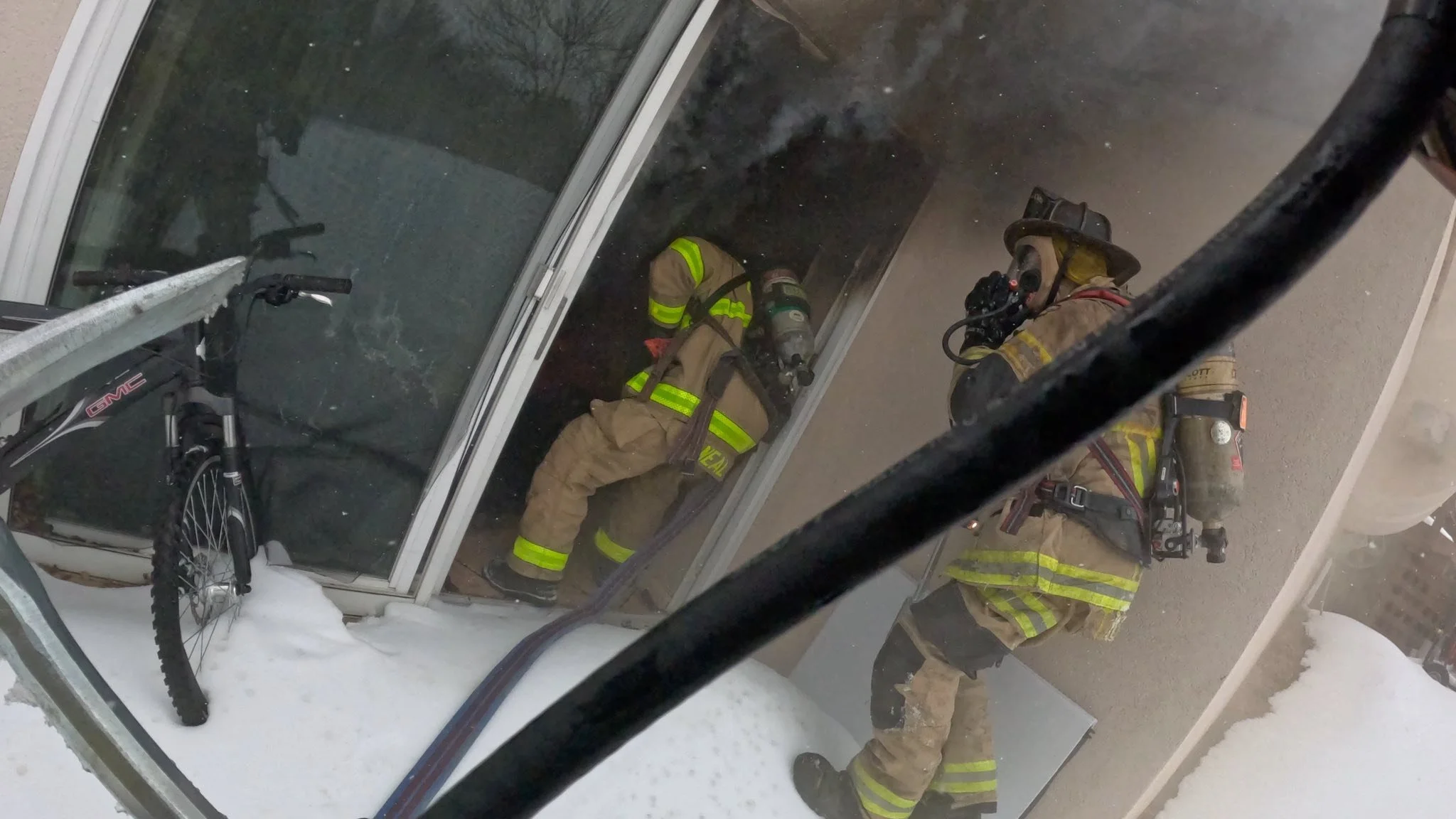 Two firefighters in full uniform and masks entering a home with smoke coming out of it. 