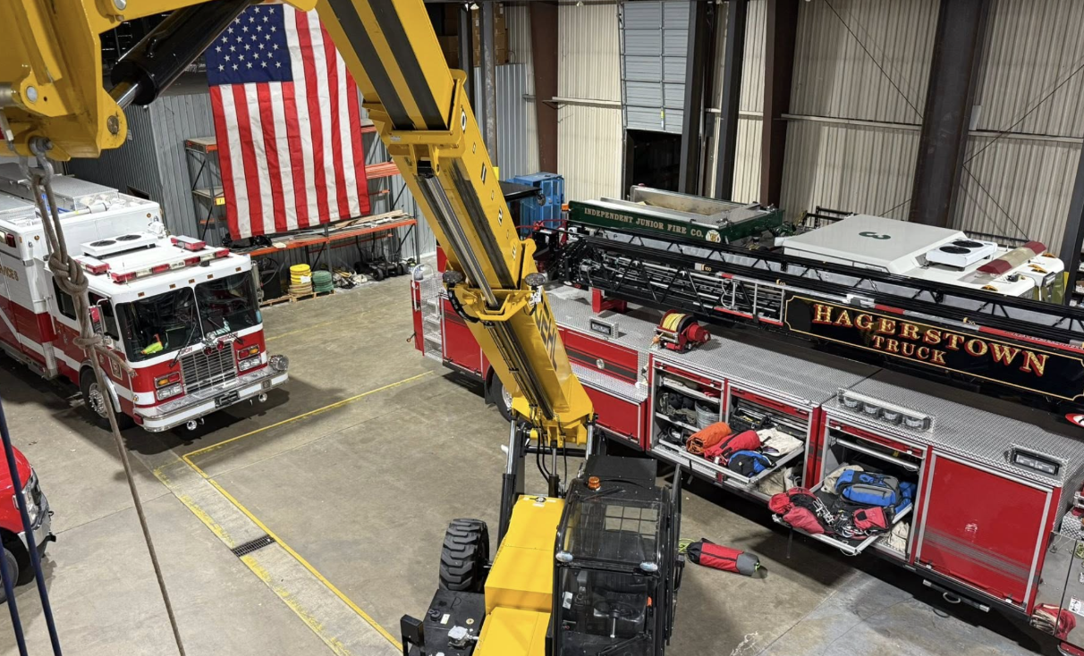 A yellow crane and two firetrucks sitting in a large building with concrete floors and an American flag hanging on the wall.