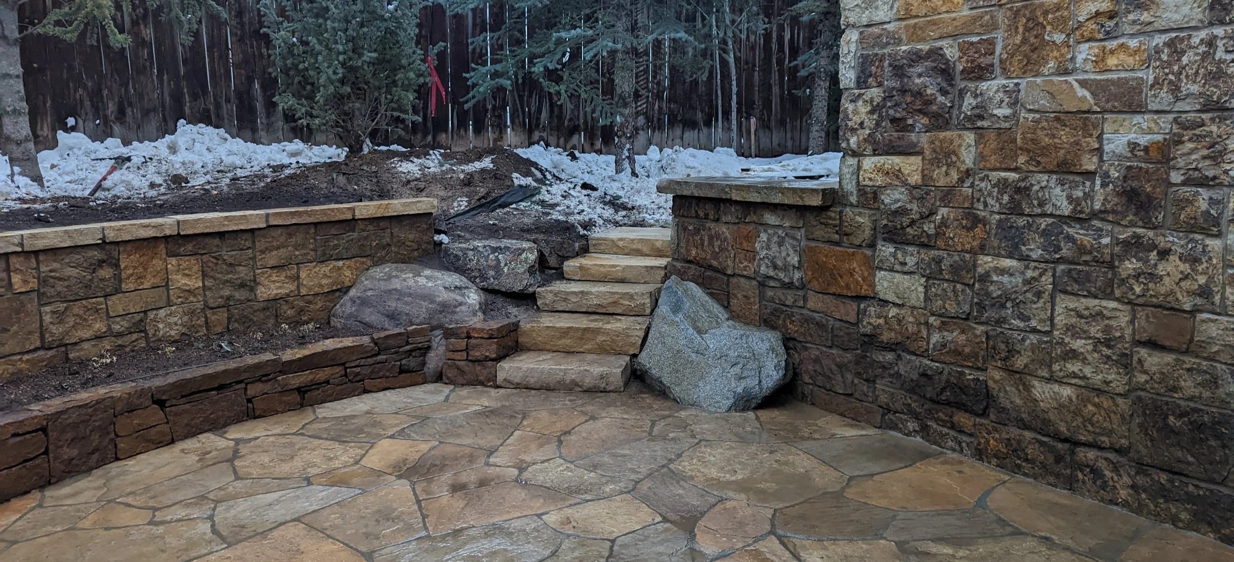 A backyard stone patio with stairs made of large stone slabs, surrounded by a stone wall on the right and a retaining wall on the left. There are large rocks near the steps and snow on the ground in the background. A wooden fence with a Christmas tree and some trees can be seen beyond the snow.