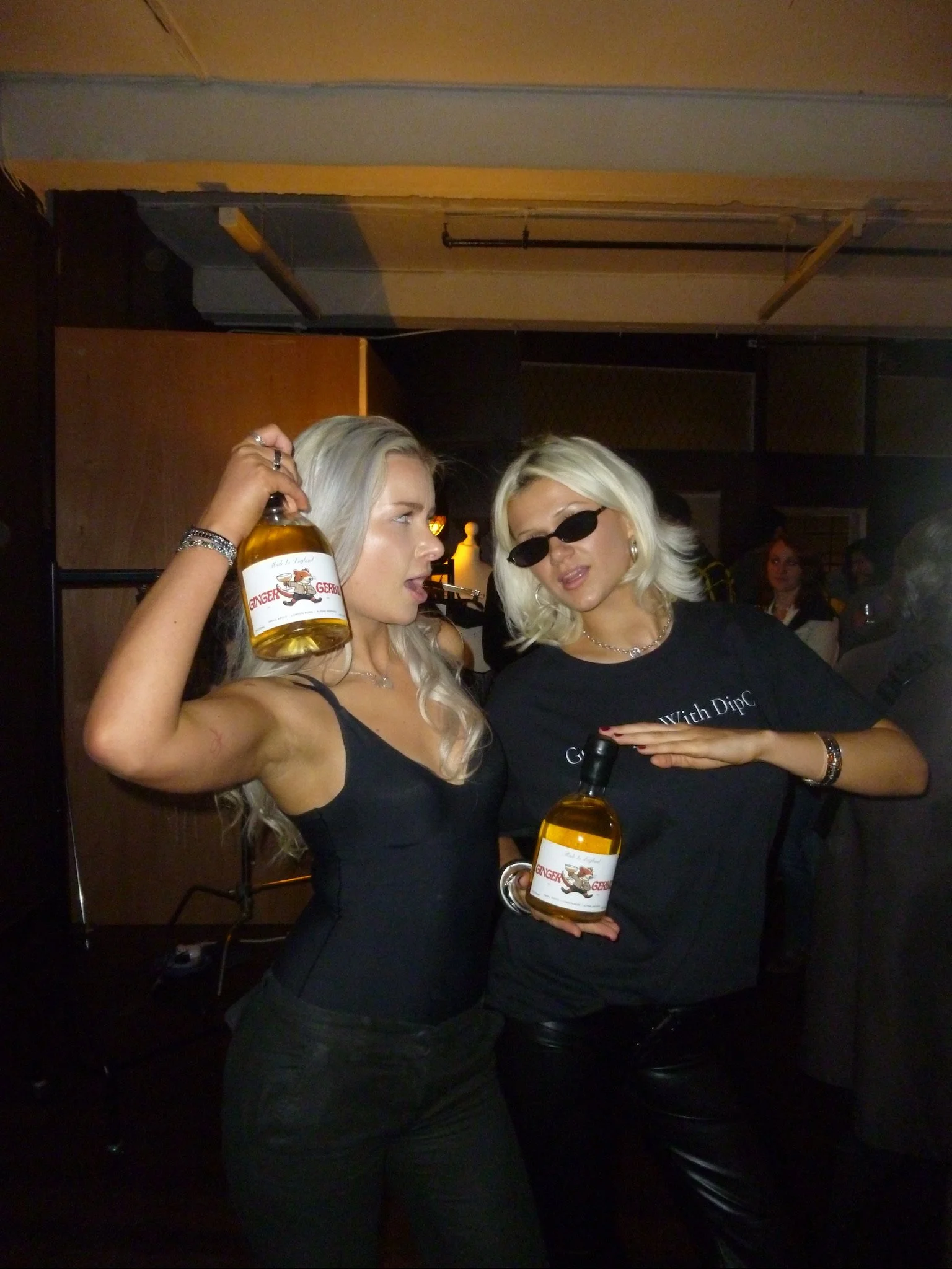 Two women holding bottles of alcoholic beverages at a social event. One woman is wearing sunglasses and a black shirt, the other is in a black outfit with long blonde hair.