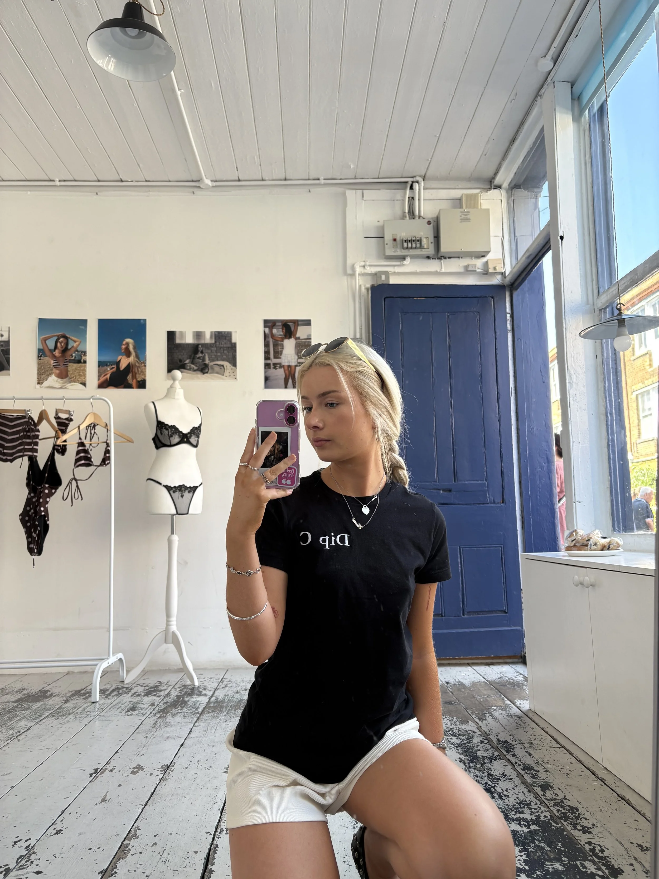 Young woman taking a mirror selfie in a store with clothing and photos on the wall, wearing a black t-shirt and white shorts.