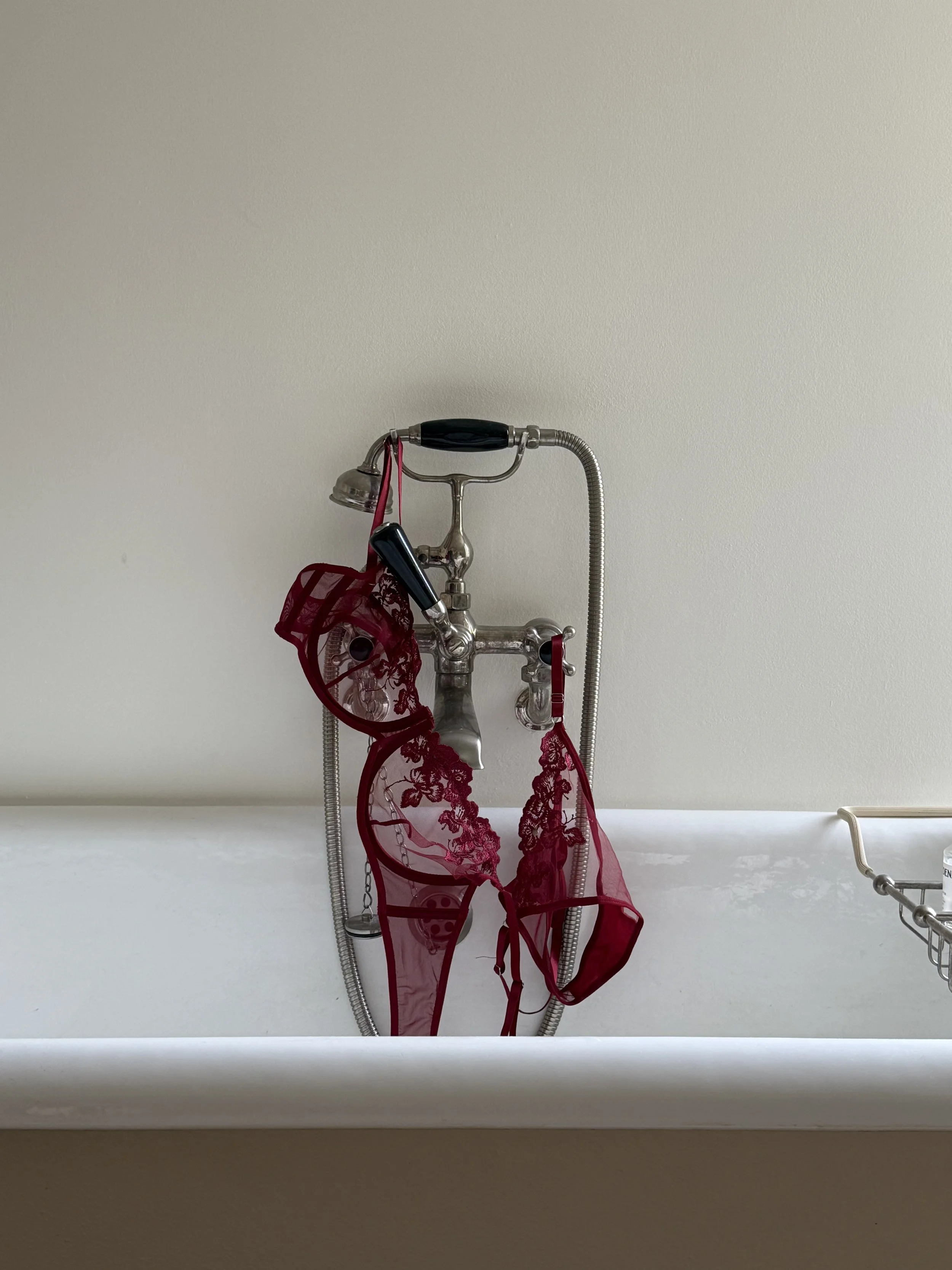 A vintage-style silver sink faucet with a handheld shower attachment on the wall above a white bathtub. Draped over the faucet is a red sheer lace lingerie set, including a bra and panties.