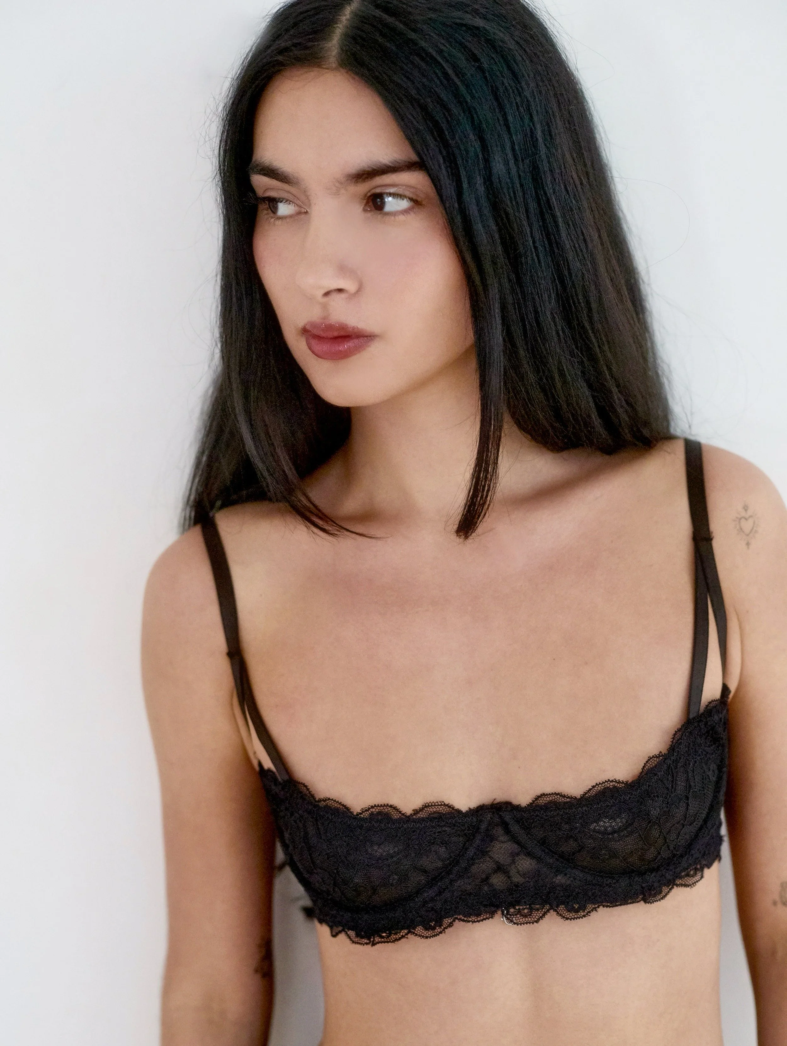 Young woman with long dark hair wearing a black lace bralette, facing slightly to the side against a plain white background.