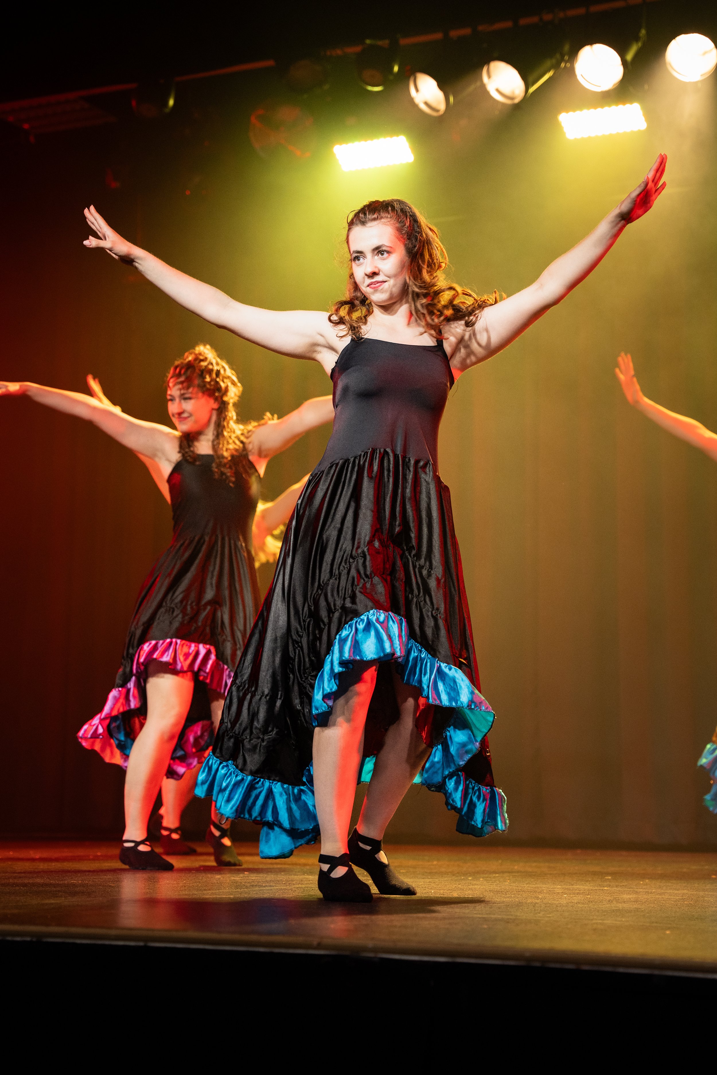 Dansers op een podium, gekleed in zwarte rokken met kleurrijke onderkant, voeren een dansroutine uit onder stageverlichting.