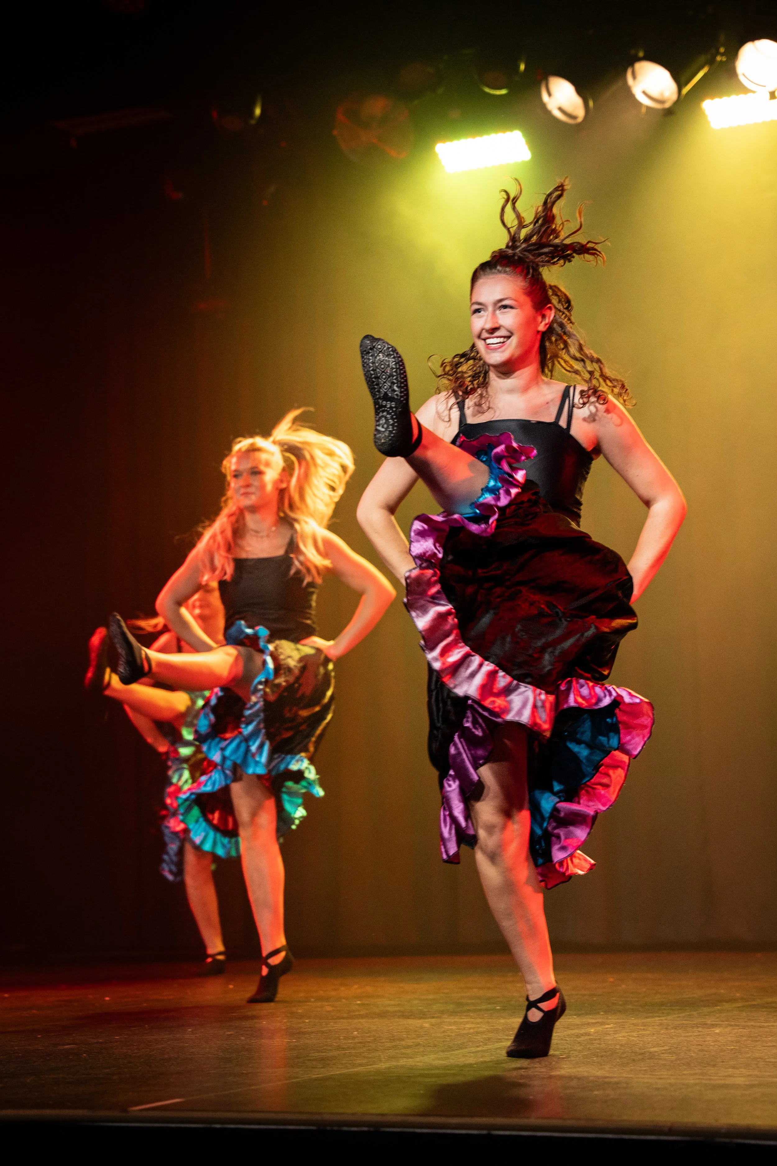 Dansende vrouwen in kleurrijke rokken en hakken tijdens een optreden op een podium met groene en gele verlichting.
