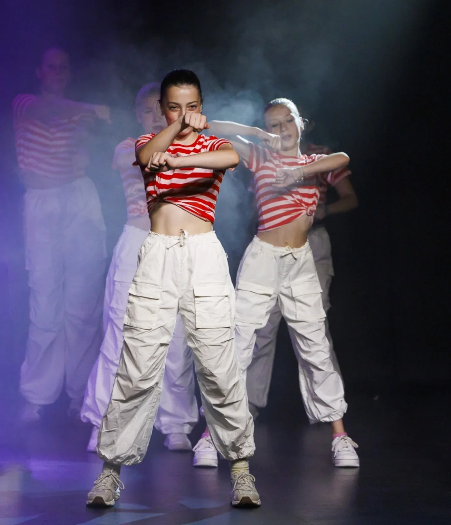 Vrouw danst op een podium, draagt een rood-wit gestreept shirt en witte broeken, met andere vrouwen op de achtergrond, in een donkere omgeving met rook en gekleurd licht.