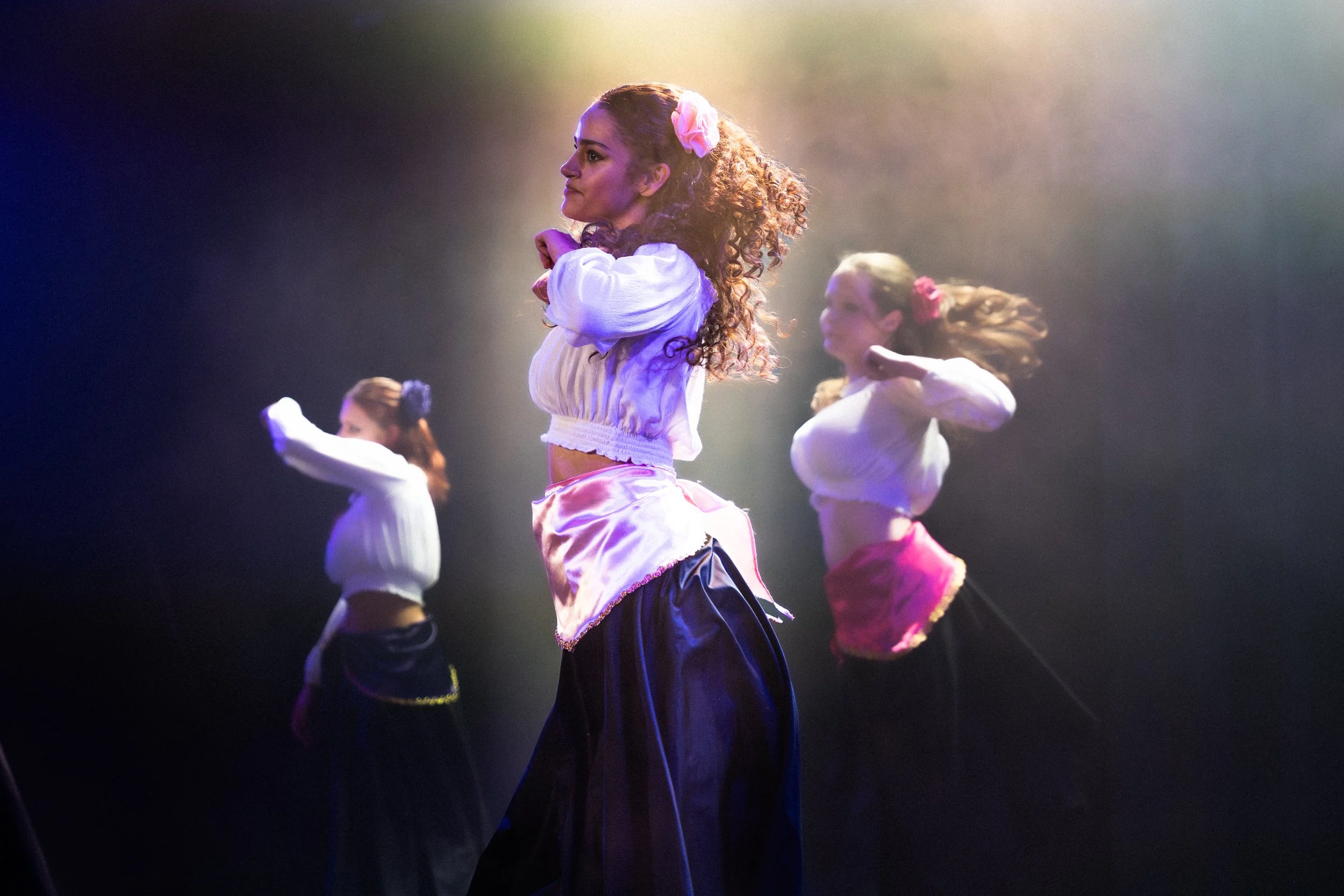 Vrouw danst op een podium met gekleurde lichteffecten, omgeven door andere dansers.