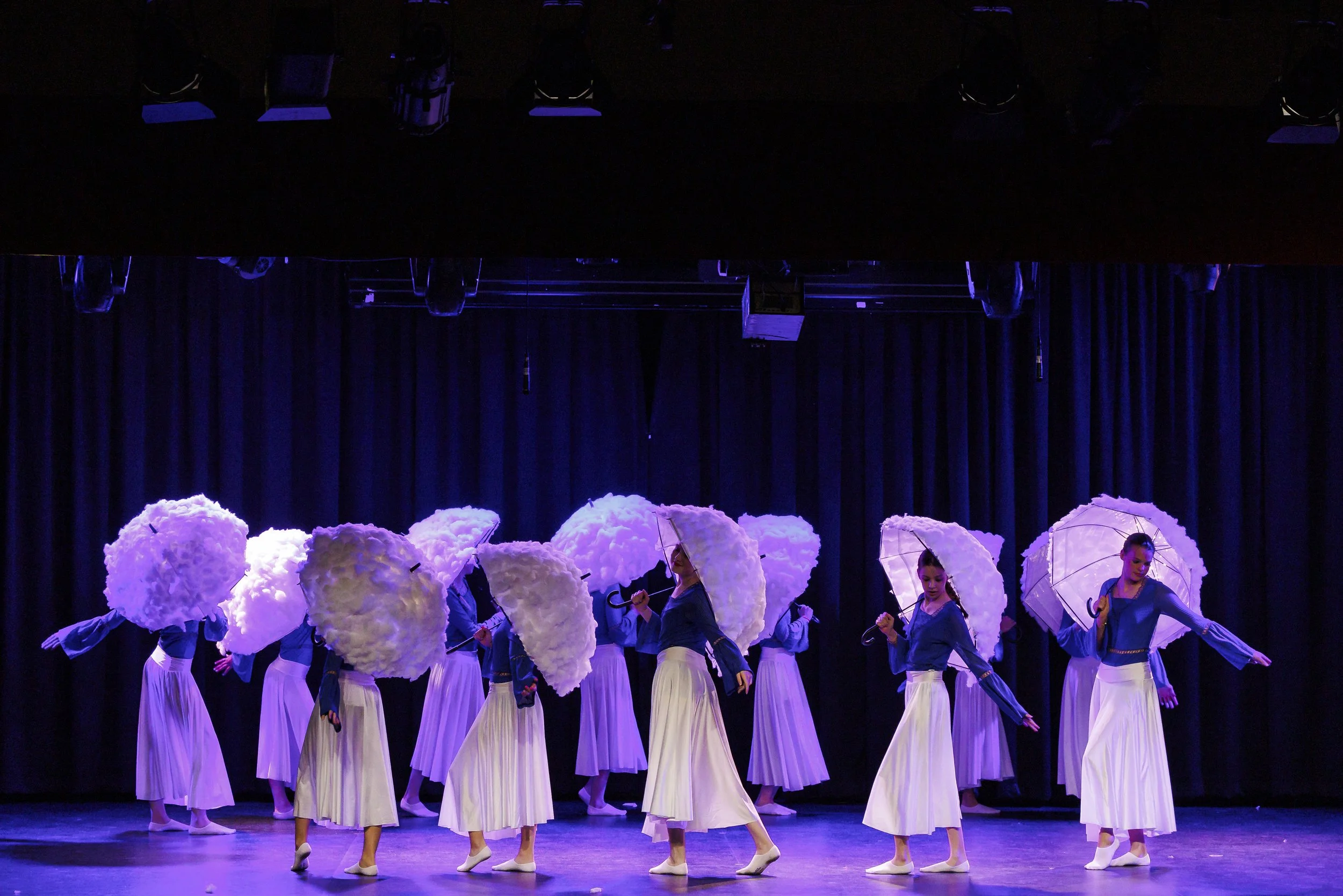 Dancers op een podium met witte roosvormige hoofddeksels en witte rokken, onder blauwe verlichting.