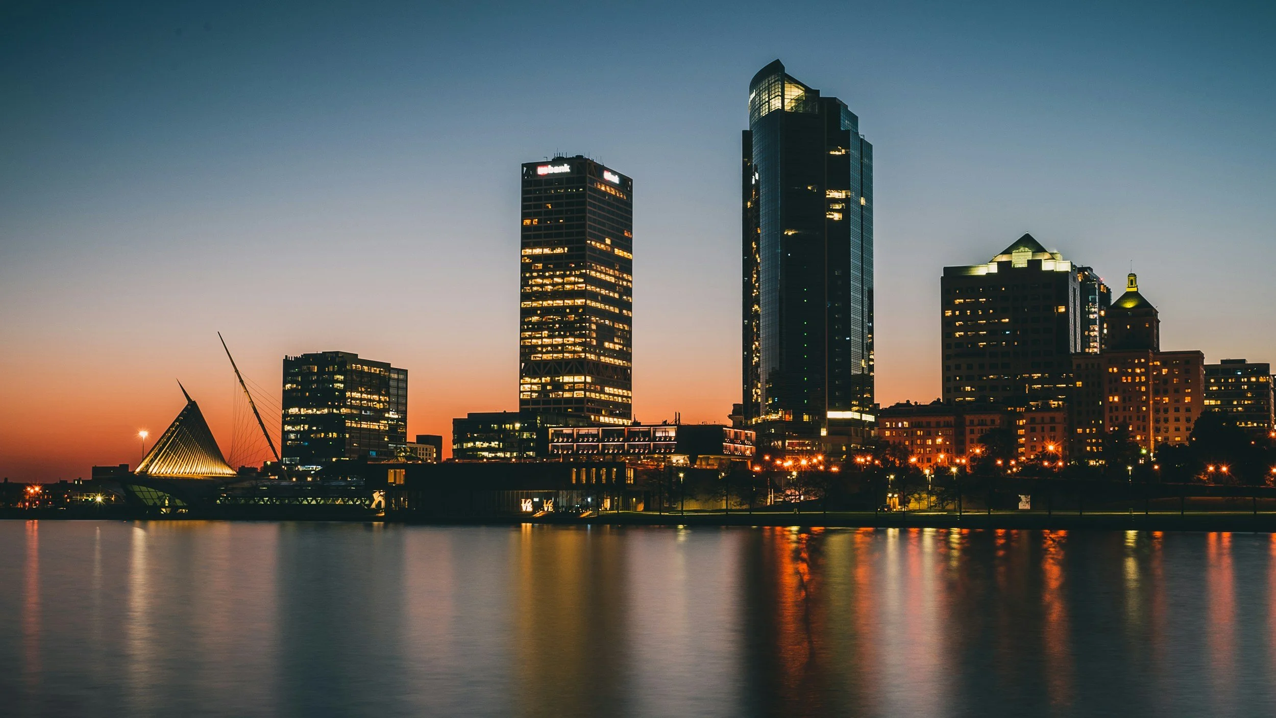 City skyline at dusk reflecting on the water, with tall modern buildings and a unique triangular-shaped structure.