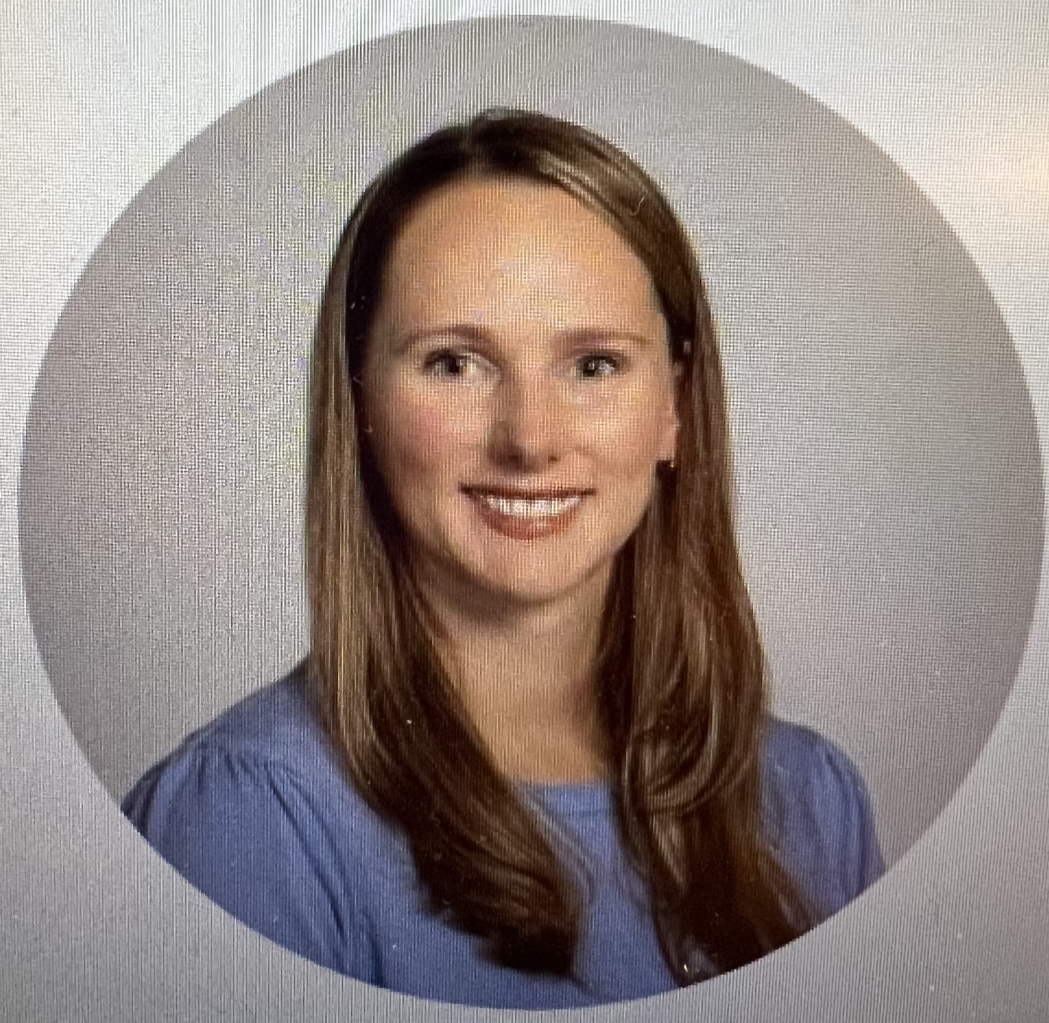 Headshot of a woman with long brown hair wearing a blue top, smiling, against a gray background.