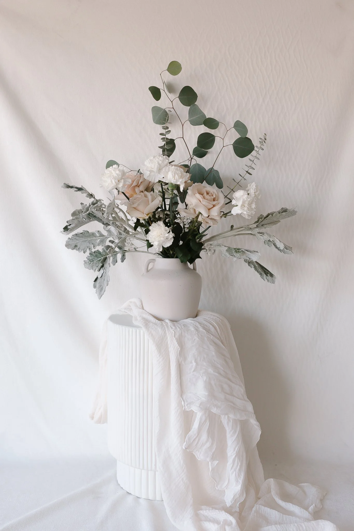 A floral arrangement in a white vase placed on a textured white pedestal with a white cloth draped over it. The arrangement includes white and pale pink roses, white carnations, and trailing eucalyptus leaves, set against a light textured wall background.