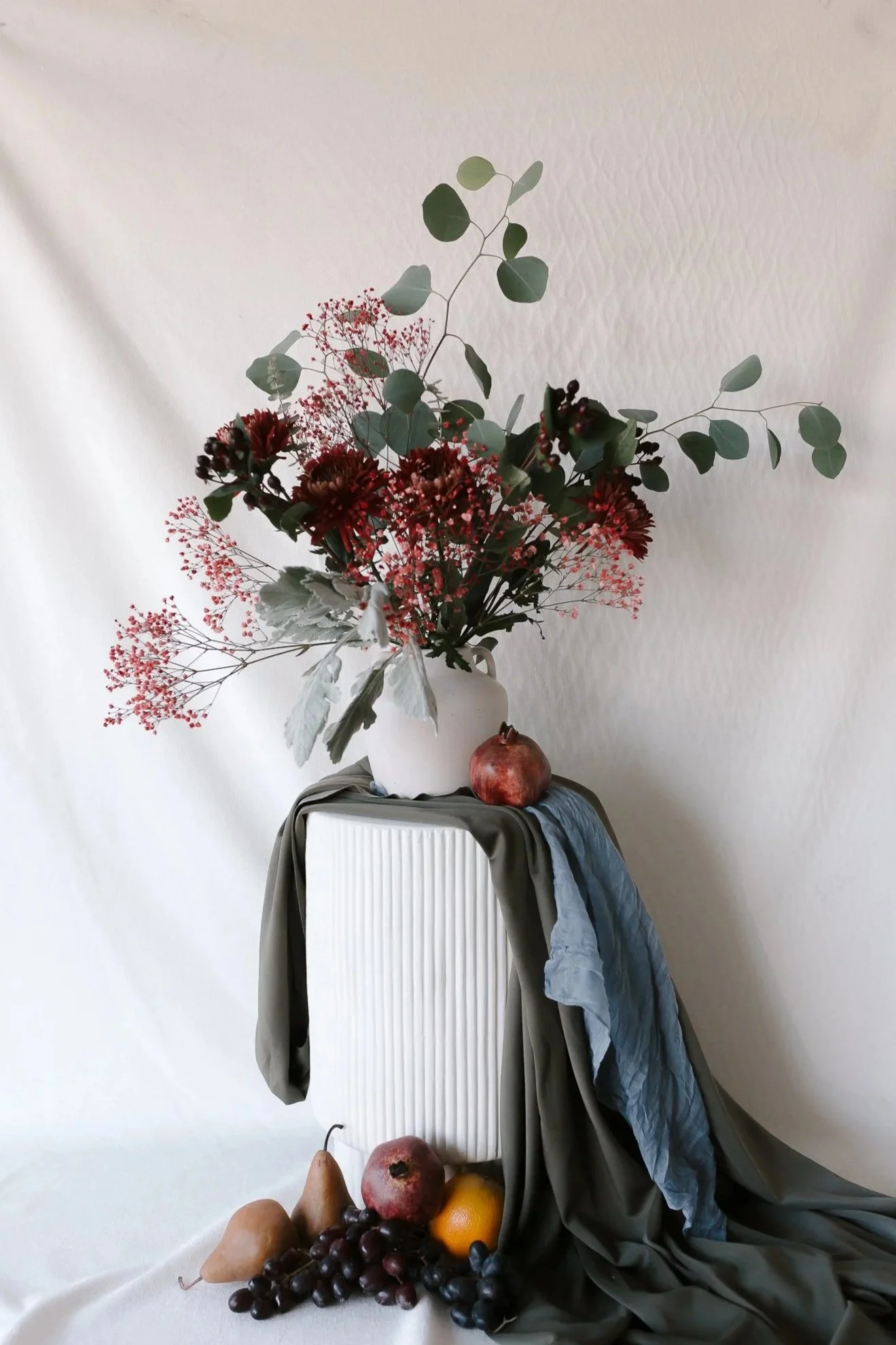 Still life composition with a large bouquet of red, pink, and green flowers and foliage in a white vase, placed on a white pedestal, topped with a pomegranate; additional fruits including pears, grapes, a lemon, and an apple are arranged on the floor and draped cloths in muted tones.