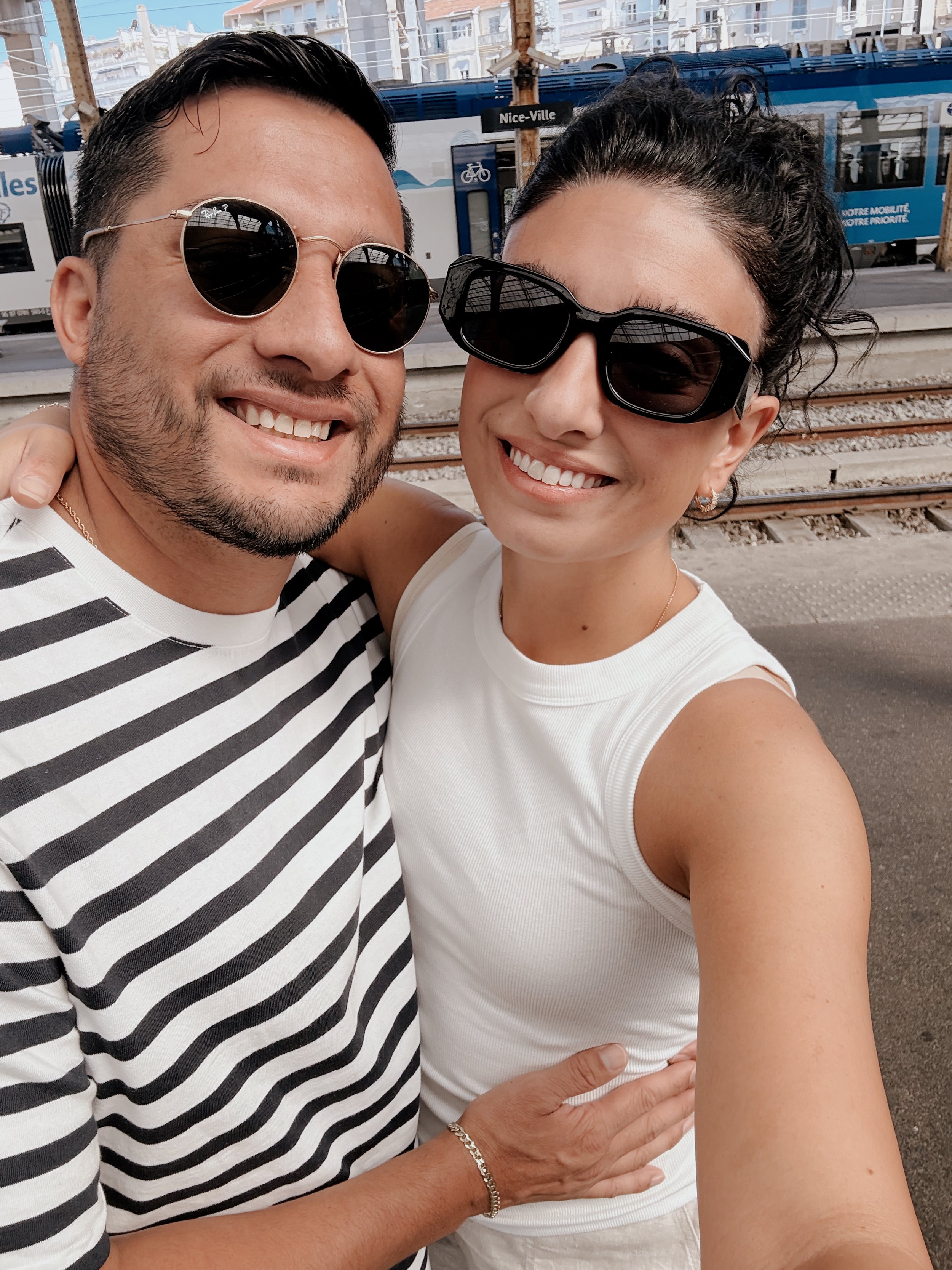A smiling man and woman taking a selfie at a train station in Nice-Ville, wearing sunglasses and casual clothes.