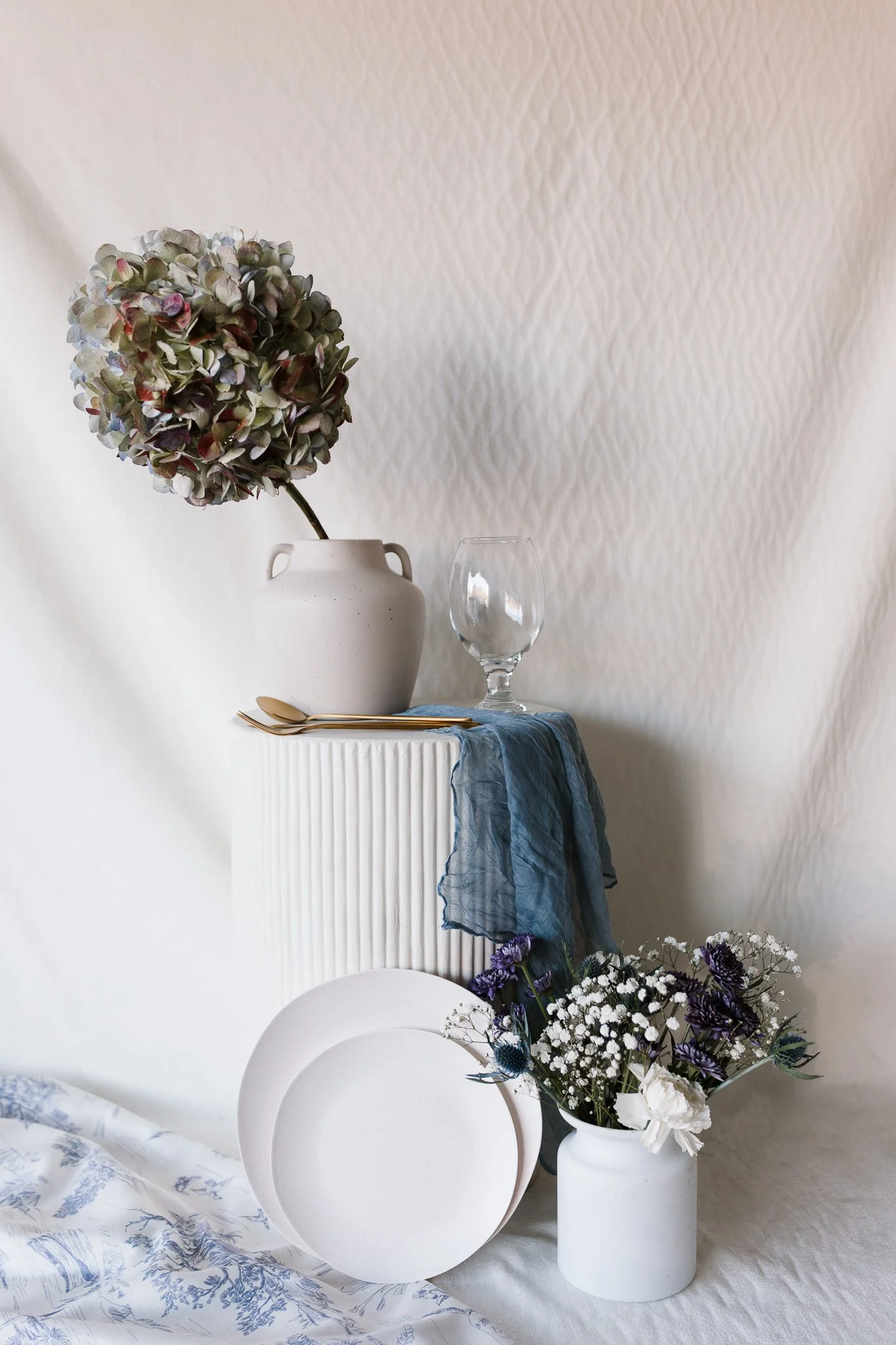 A minimalist still life with a white vase holding a pink and green hydrangea, a clear wine glass, a gold spoon and fork, a blue cloth, white plates leaning against a white textured background, and a white vase with purple and white flowers, all arranged on a light fabric surface.