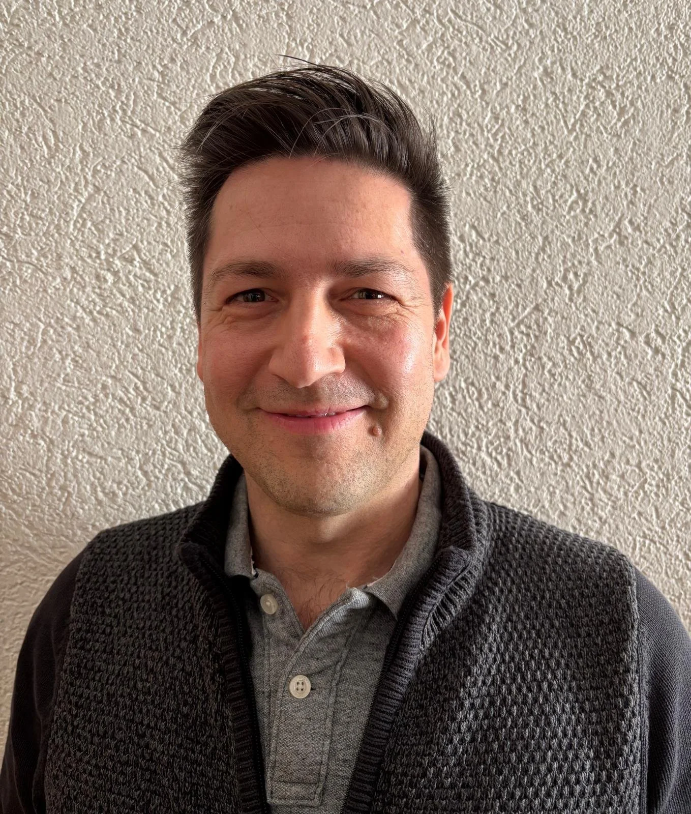 Ein lachender Mann mit dunklem Haar vor einer strukturierten Wand.