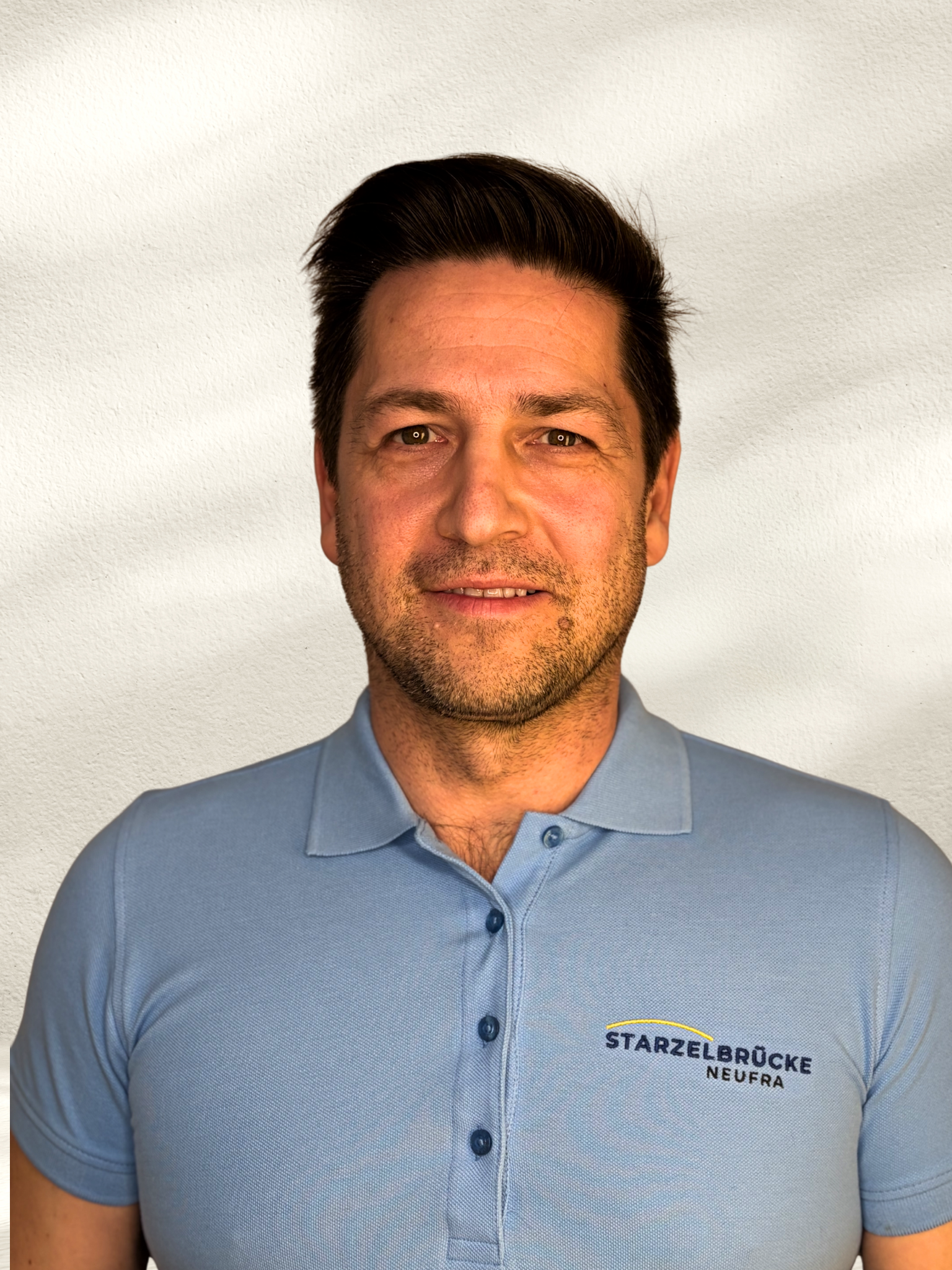 Ein lachender Mann mit dunklem Haar vor einer strukturierten Wand.