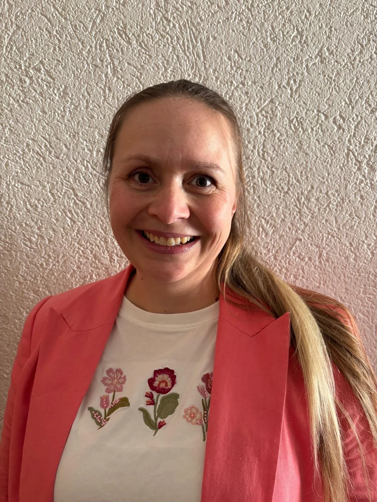 Fröhliche Frau mit braunen Haaren, blass und in einem rosa Blazer, vor einer hellen, strukturierten Wand.