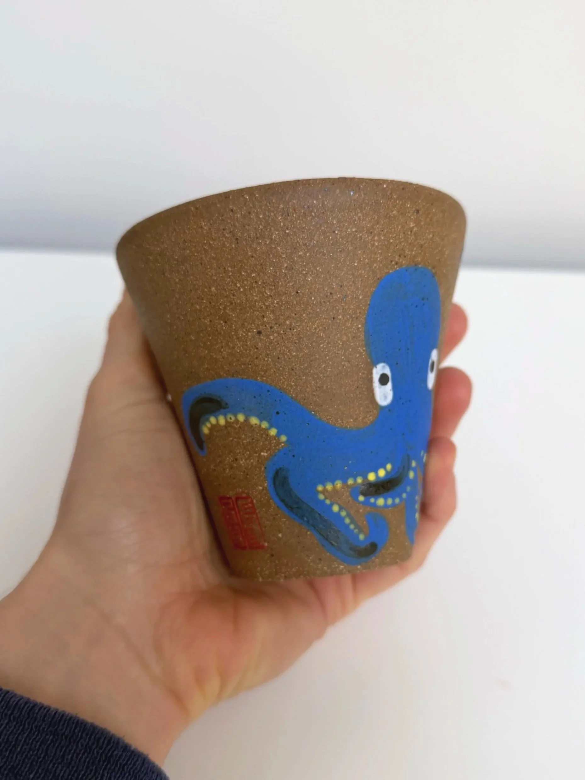 Two ceramic cups with black monkey illustrations and red faces, held in a person's hand against a wooden background.