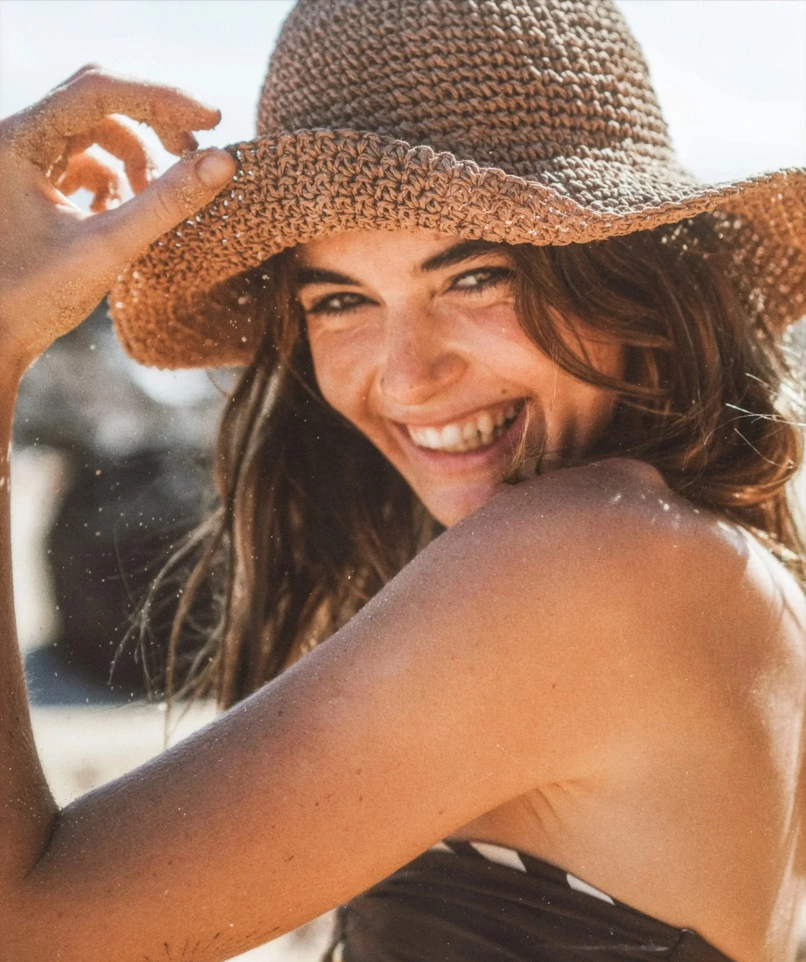 A woman with brown hair smiling, wearing a wide-brimmed straw hat, at the beach in sunlight.