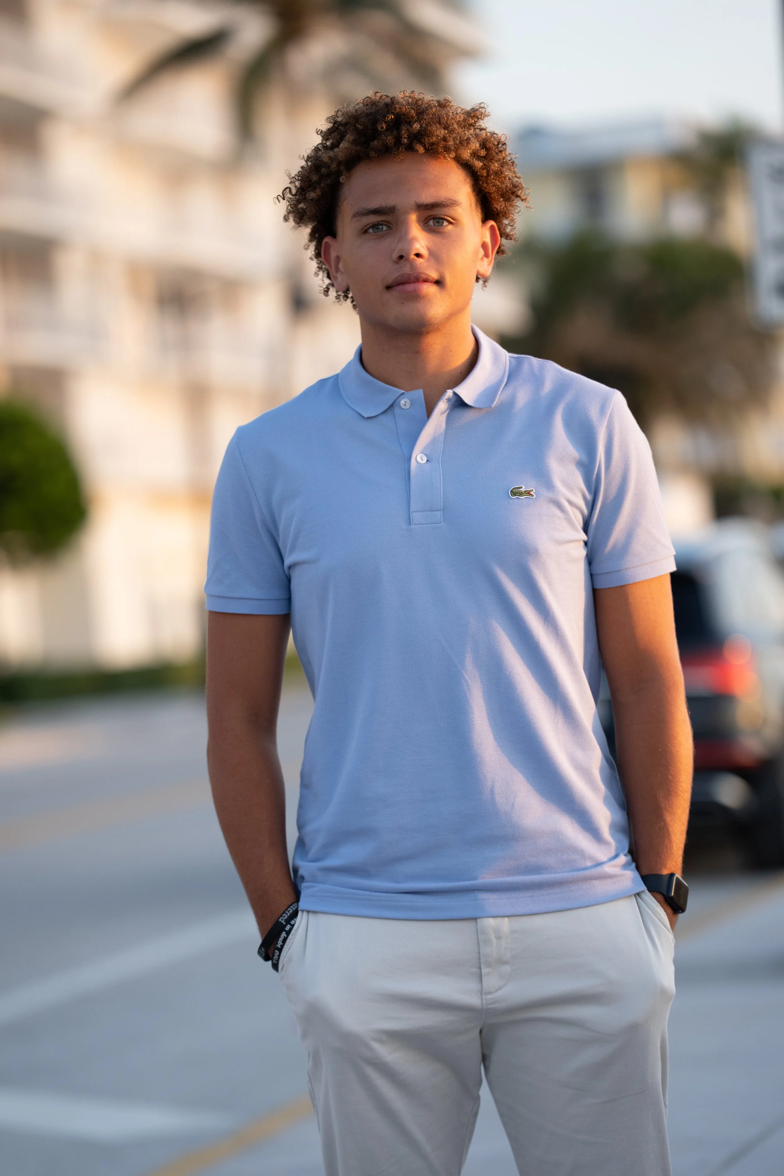 A young man with curly hair wearing a light blue polo shirt and white pants, standing outdoors in a blurred street scene during daylight.