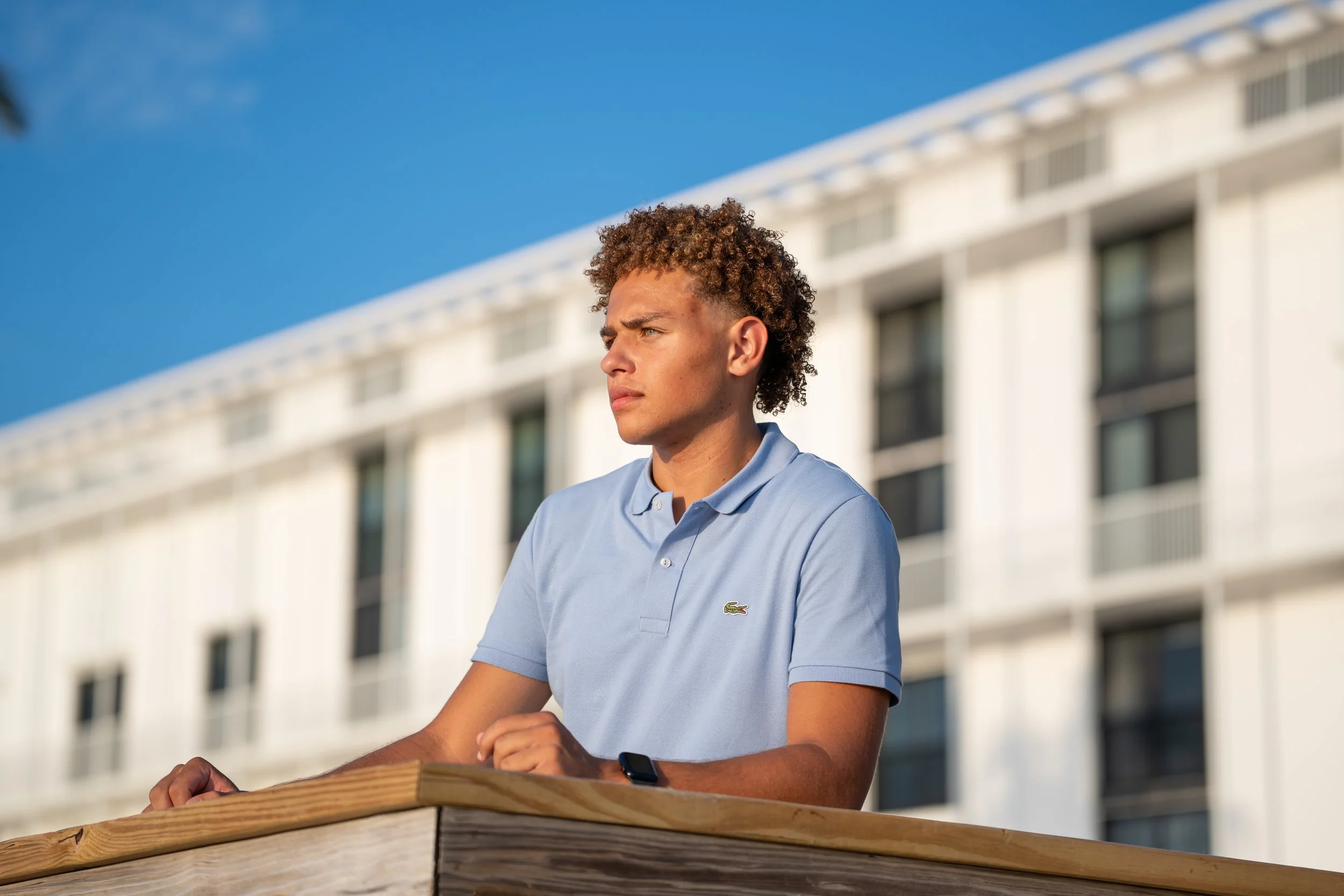 A young man with curly hair wearing a light blue polo shirt and smartwatch sitting outdoors at a wooden surface, with a modern building and a blue sky in the background.
