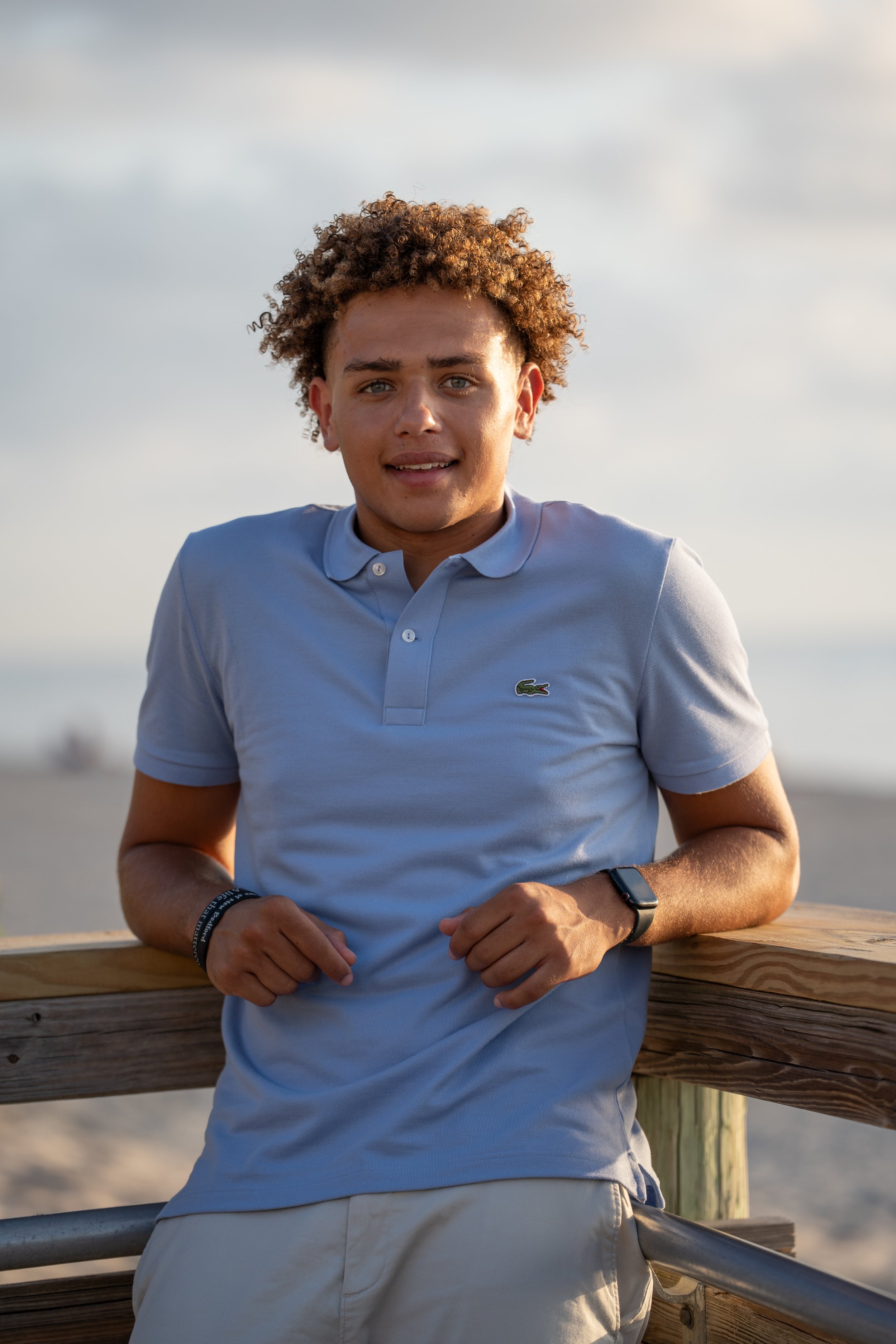 A young man with curly hair wearing a light blue Lacoste polo shirt and beige shorts standing outdoors on a wooden pier or railing, with a cloudy sky and ocean in the background.