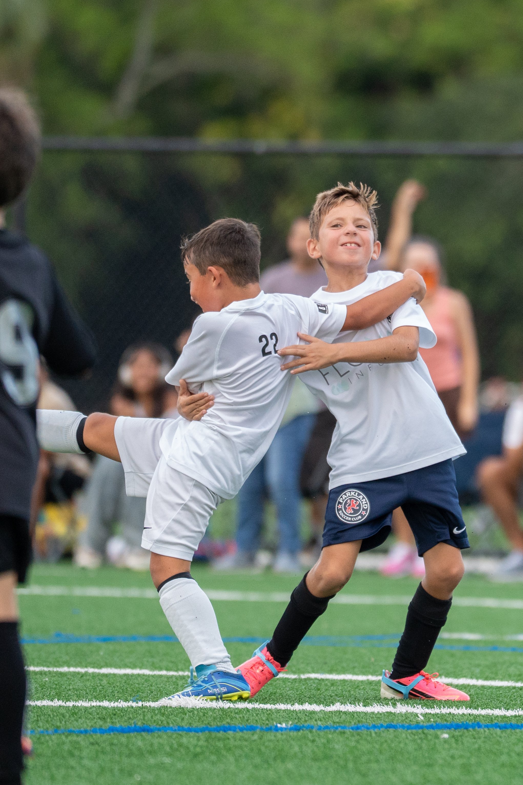 Two young boys playing soccer on a field, embracing each other in celebration. One boy is wearing a white jersey with the number 22, and the other is in a navy blue uniform with a logo that reads 'Parkland Soccer Club.'