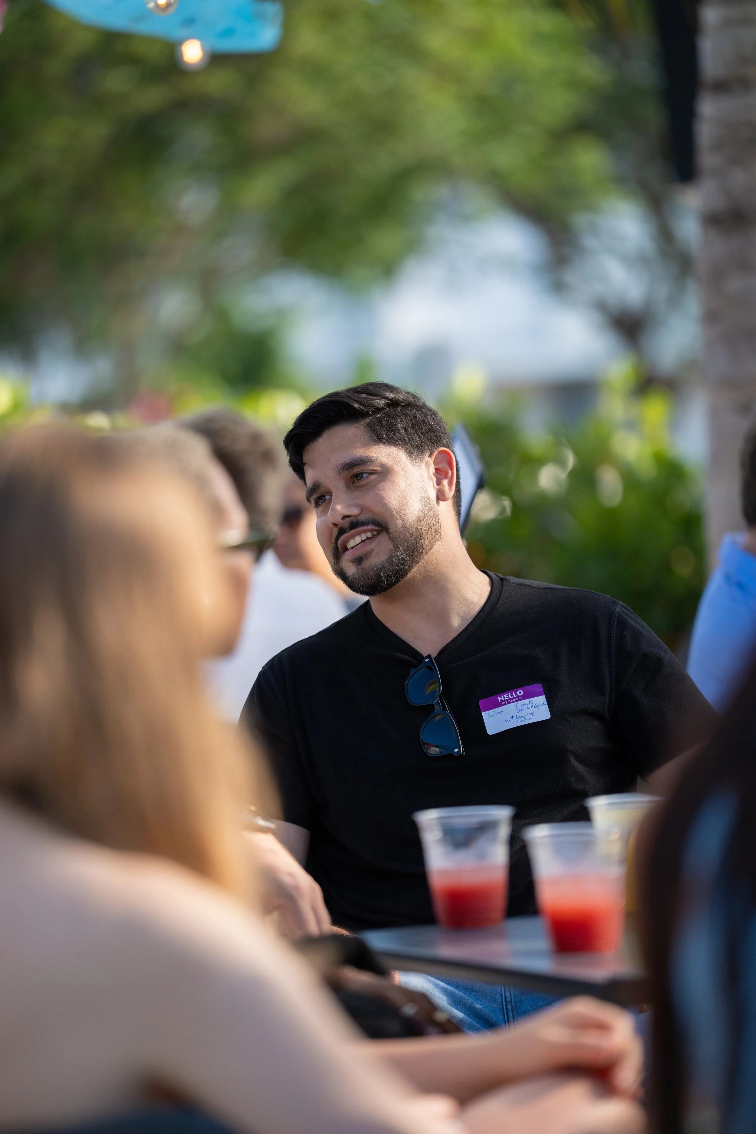 A man with a dark beard and hair, wearing a black shirt with sunglasses hanging from the collar, is smiling and talking to a group of people at an outdoor gathering. He is wearing a name tag. The background has trees and blurred people, with sunlight