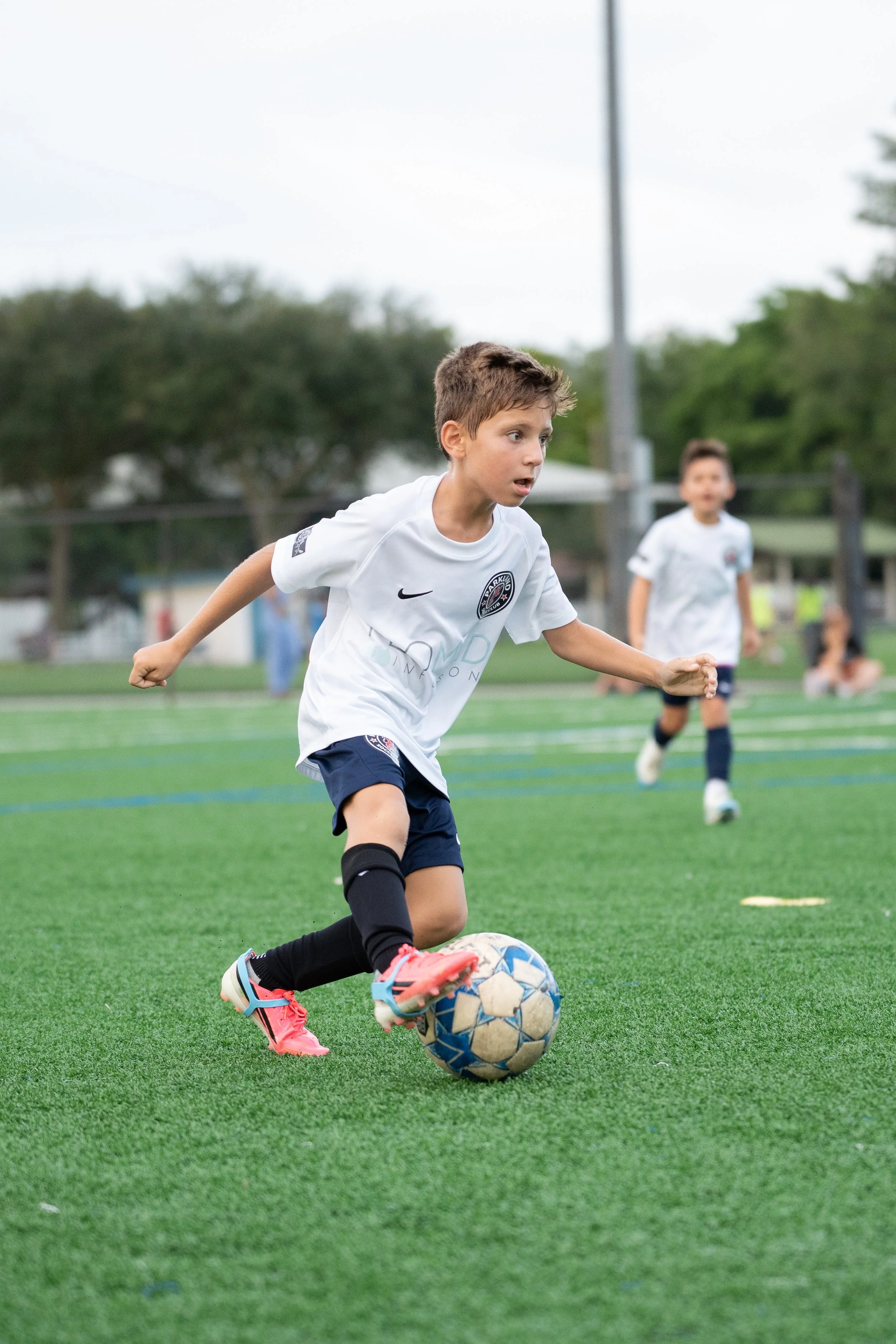 A young boy in a white soccer jersey and black shorts is actively playing soccer on a green field, dribbling a blue and white ball with his right foot, with other children and trees in the background.