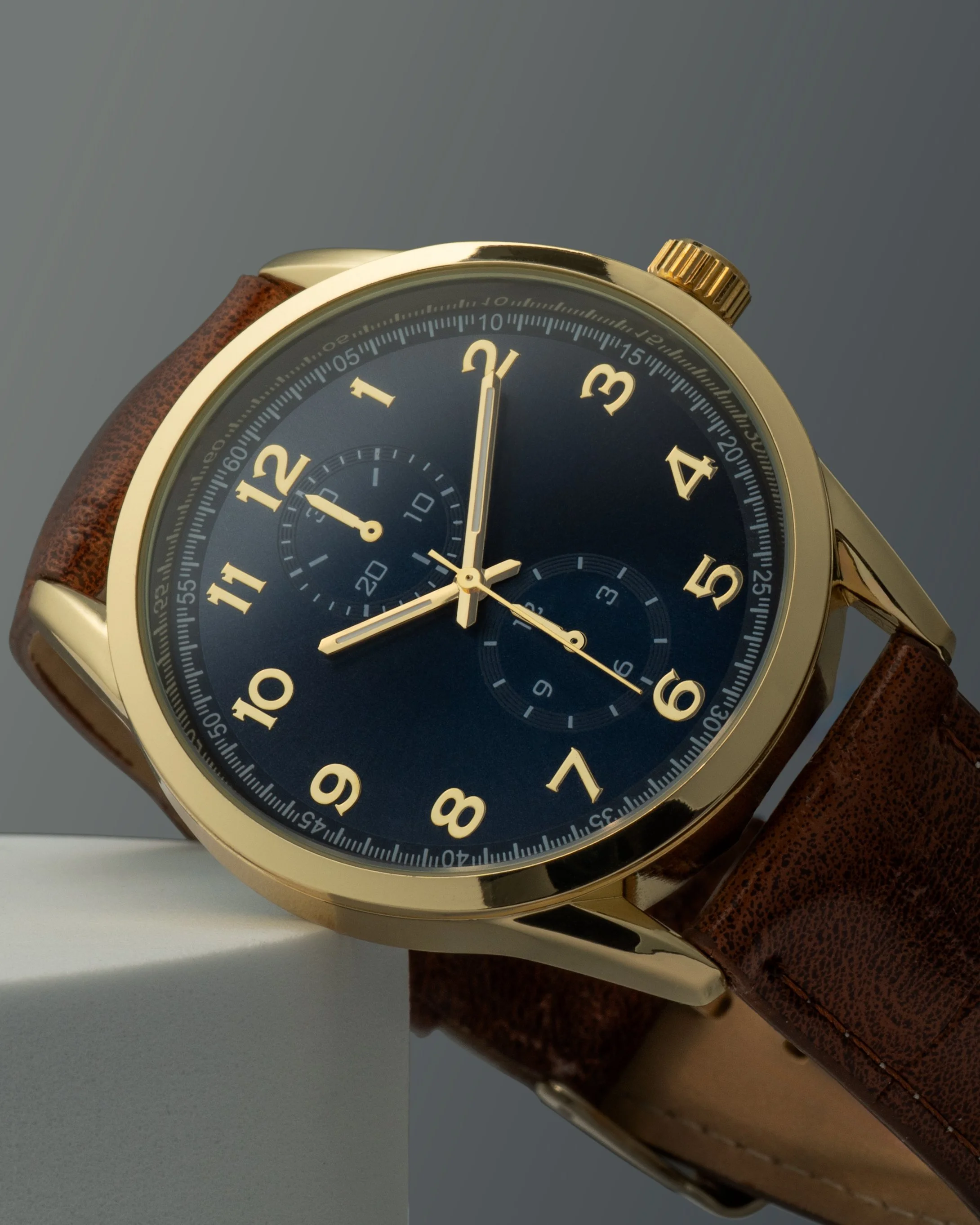 Close-up of a stylish wristwatch with a gold casing, a dark blue dial, and a brown leather strap, showing the time at 12:00.