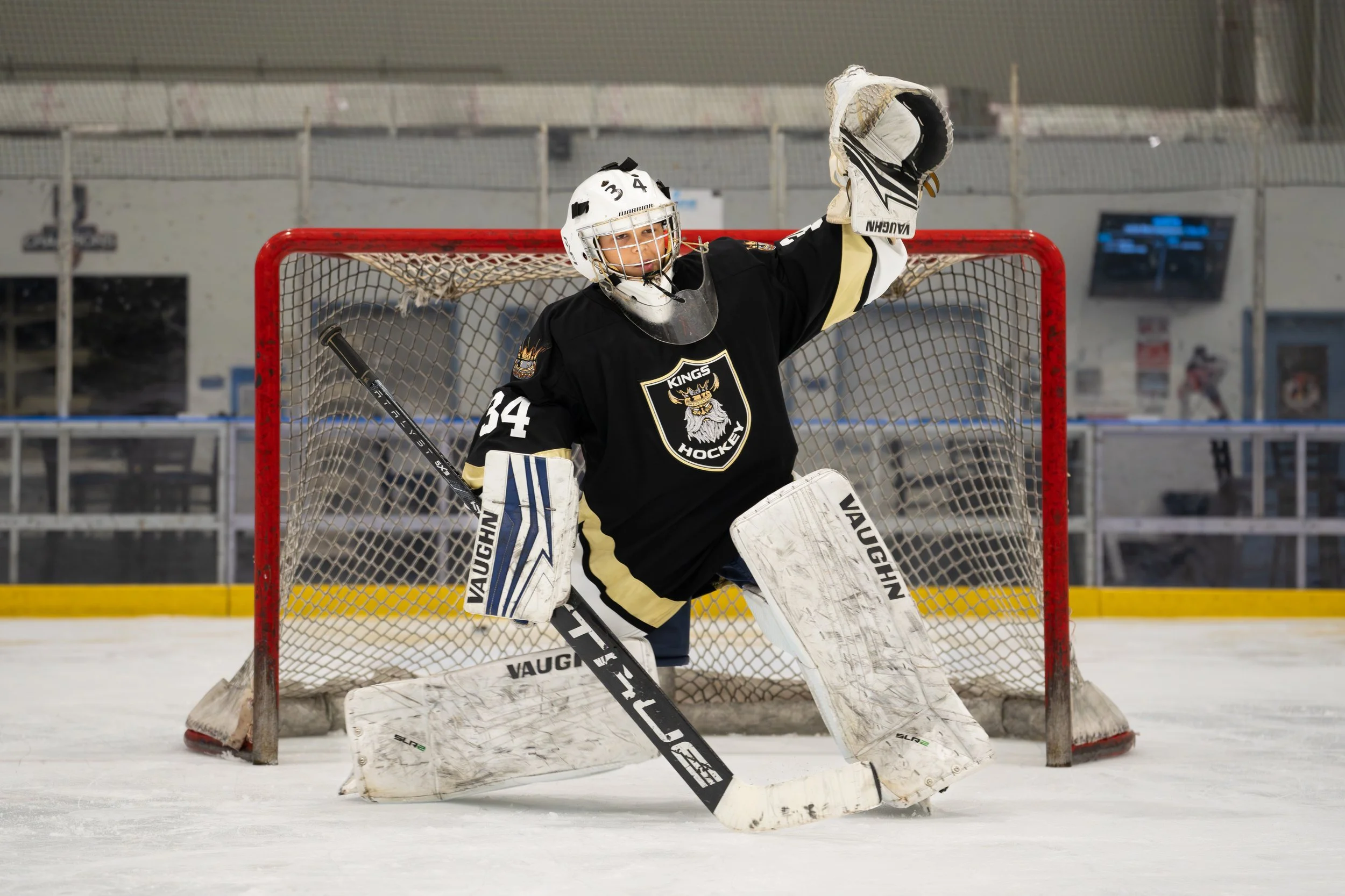 A hockey goaltender wearing black and gold gear, standing in front of the goal, ready to block a shot during a game at an indoor ice rink.