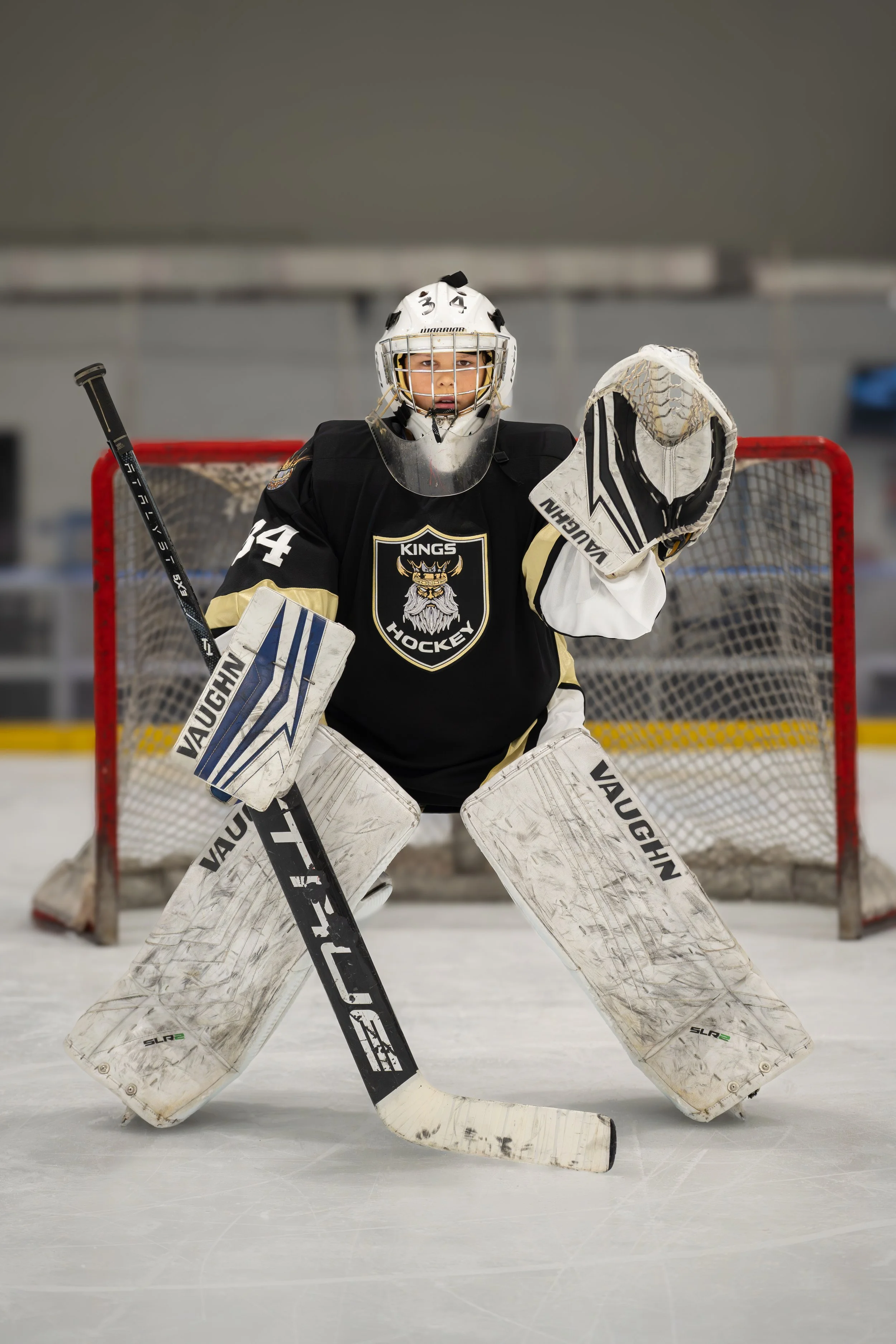 A young ice hockey goalie stands in front of the net on the ice rink, wearing a black jersey with the Kings Hockey logo, helmet, gloves, and padded leg guards, holding a hockey stick.