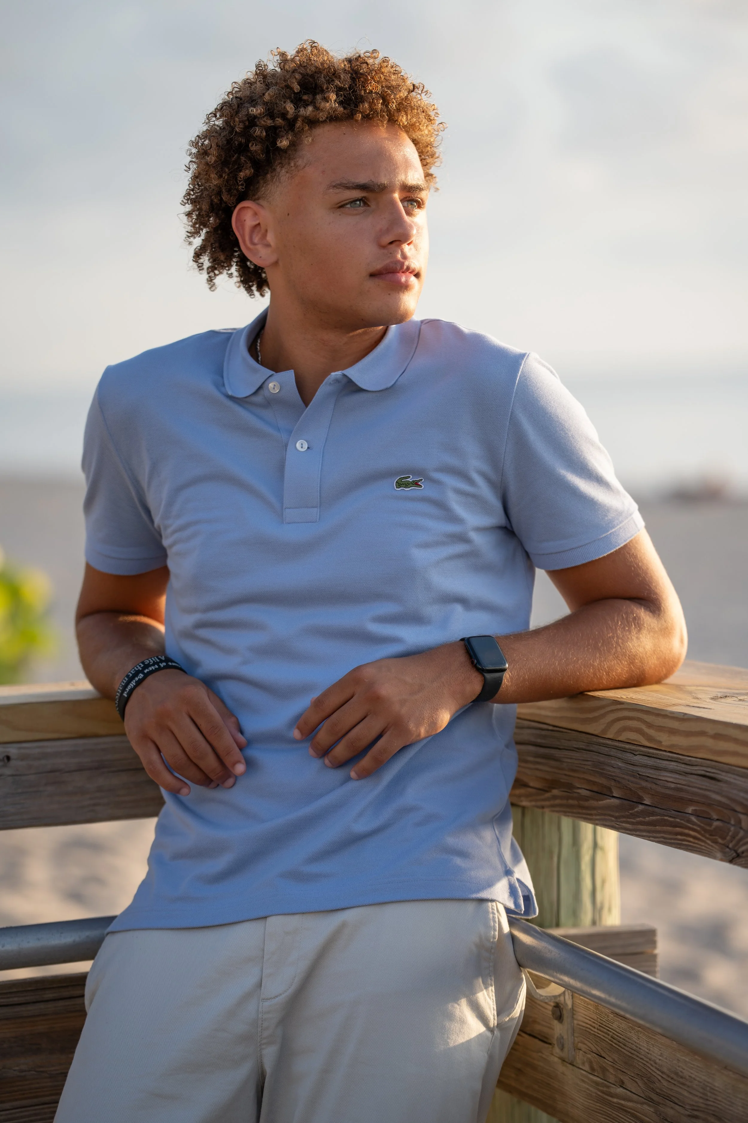 Young man with curly hair wearing a light blue polo shirt and khaki shorts, leaning on a wooden railing outdoors, looking into the distance with a thoughtful expression, during sunset.