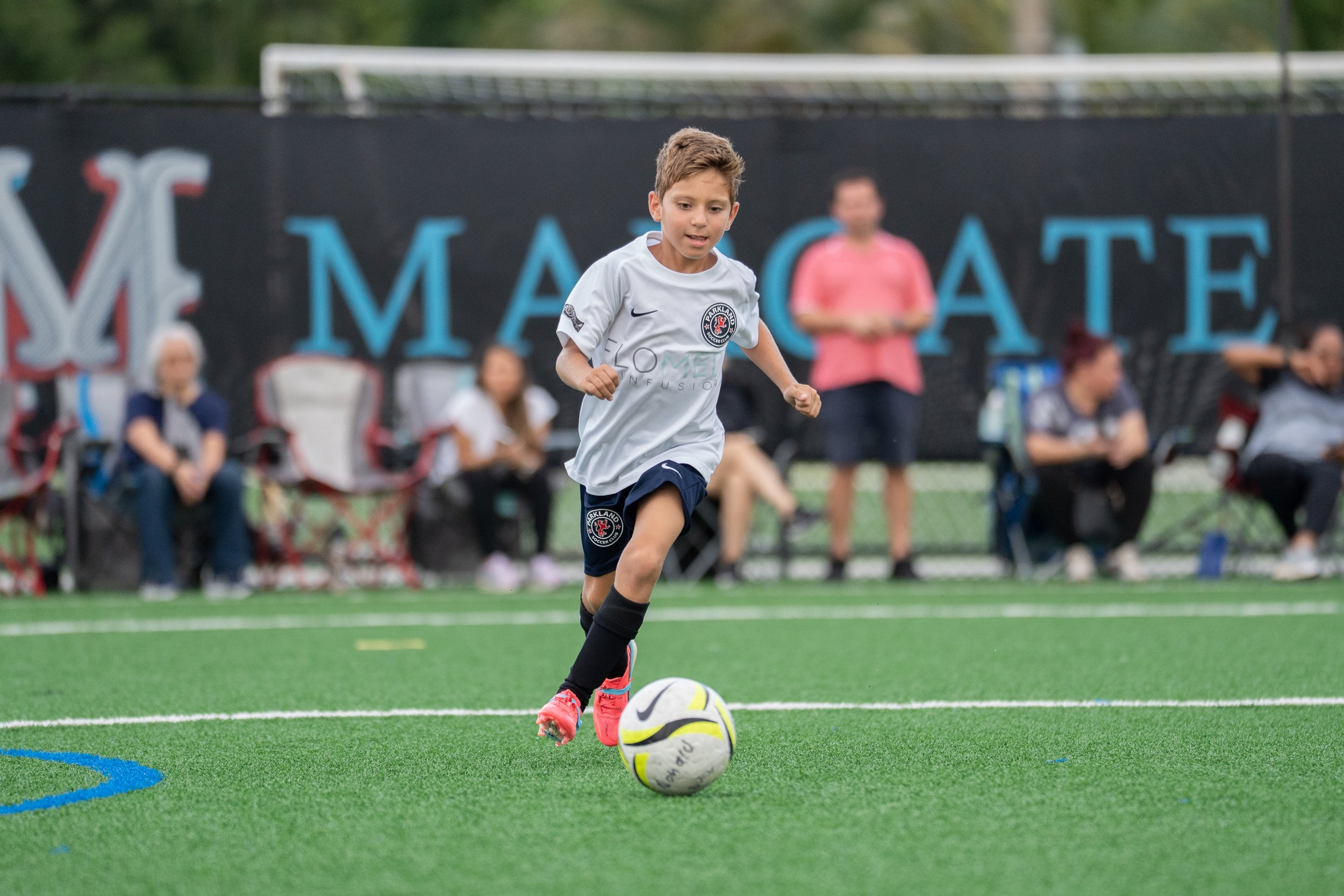 A young boy in a white soccer jersey and black shorts runs on a soccer field, dribbling a soccer ball with yellow, black, and white coloring. In the background, spectators are seated and standing near a black fence with colorful advertising banners.