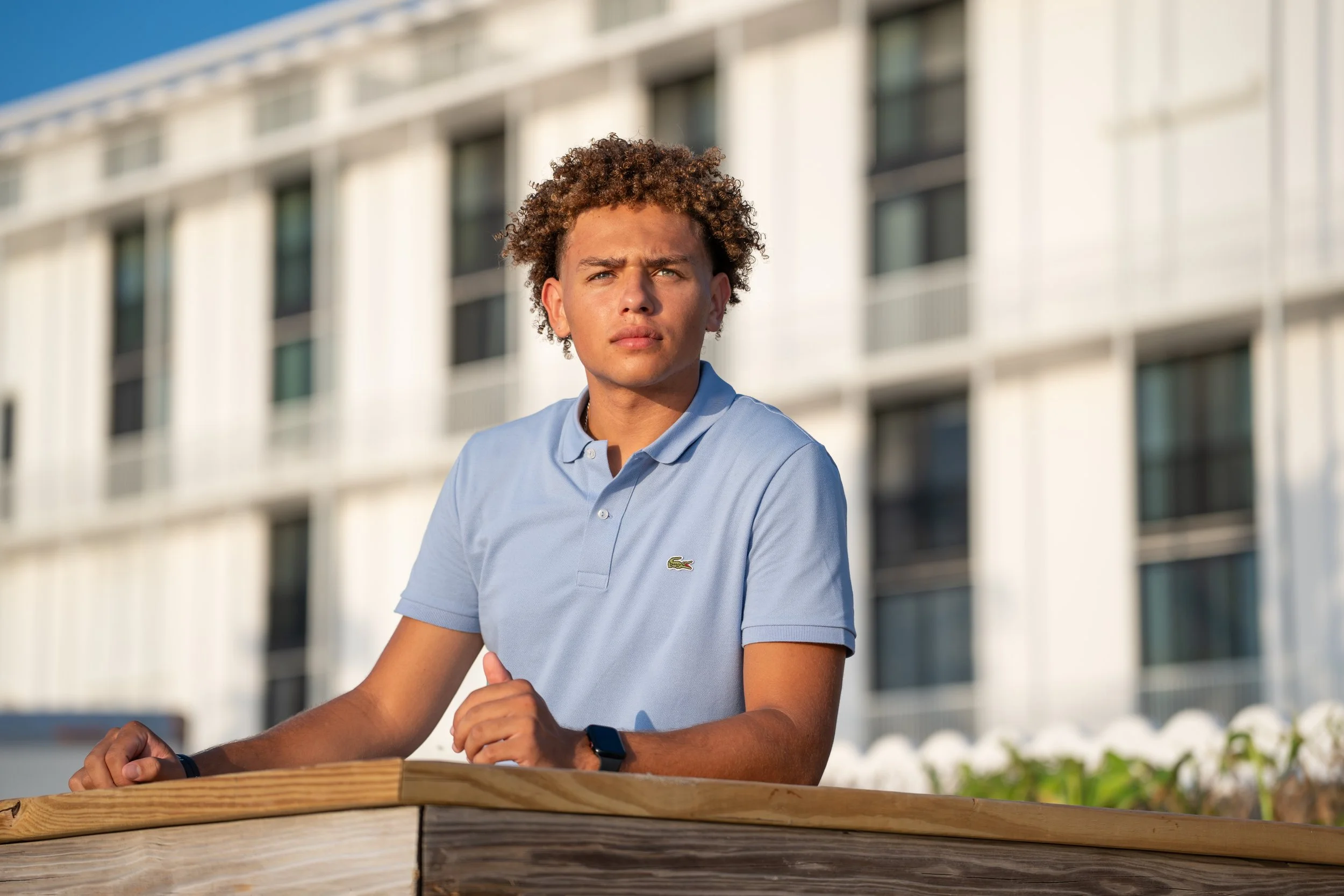 Young man with curly hair wearing a light blue Lacoste polo shirt, standing outside in front of a modern building with glass windows, looking into the distance.
