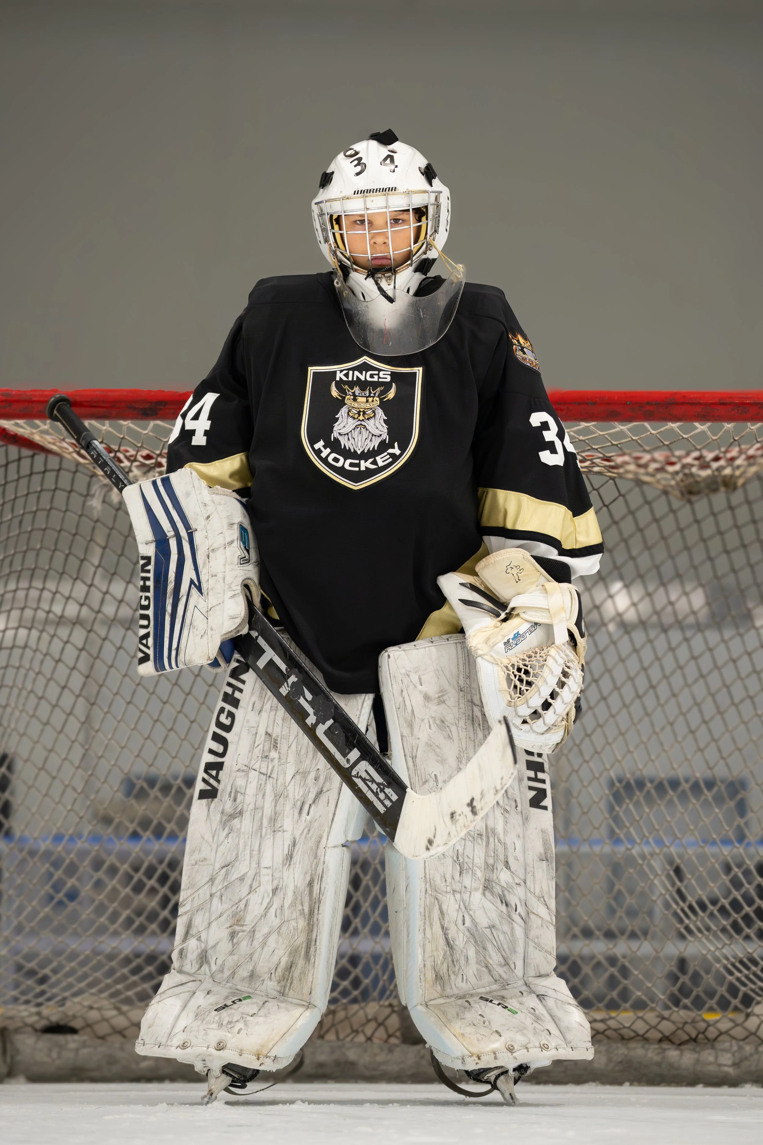 Ice hockey goalie standing in front of the goal net on ice rink, wearing black jersey with a lion logo, white leg pads, and helmet with face cage.