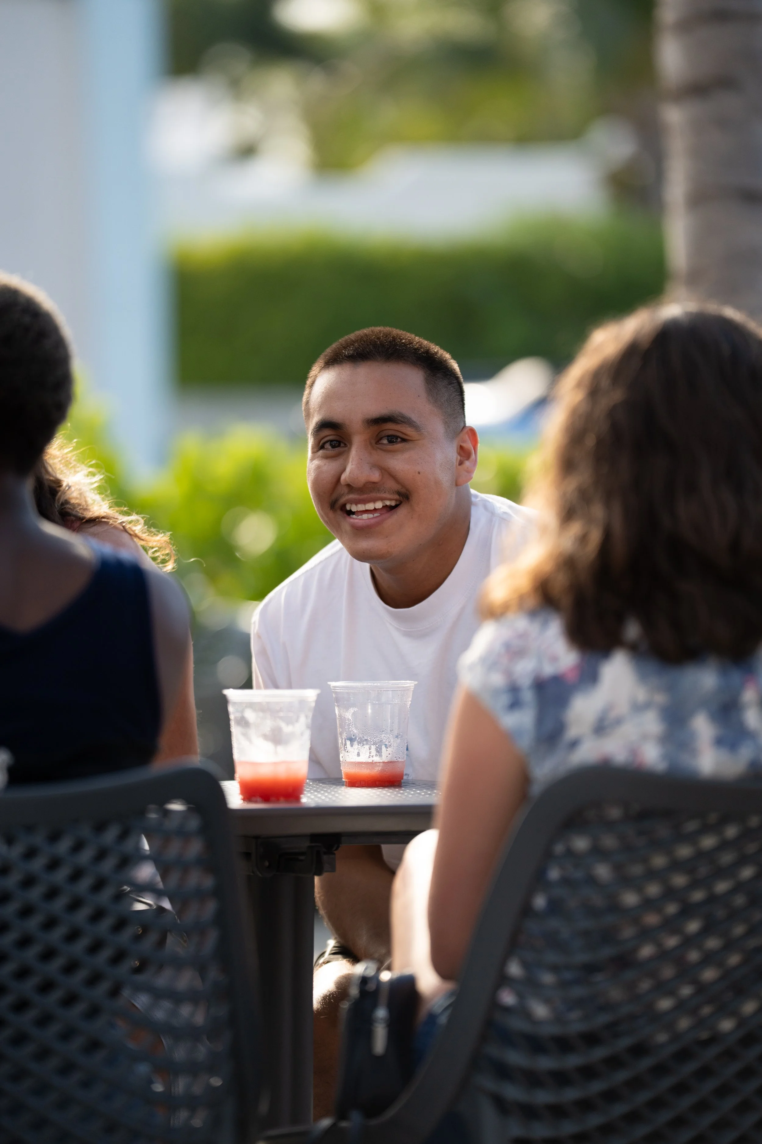 A young man with short hair smiling and talking to two women during an outdoor gathering, glasses of beverage in front of him, set against a background of greenery and a blurred outdoor setting.