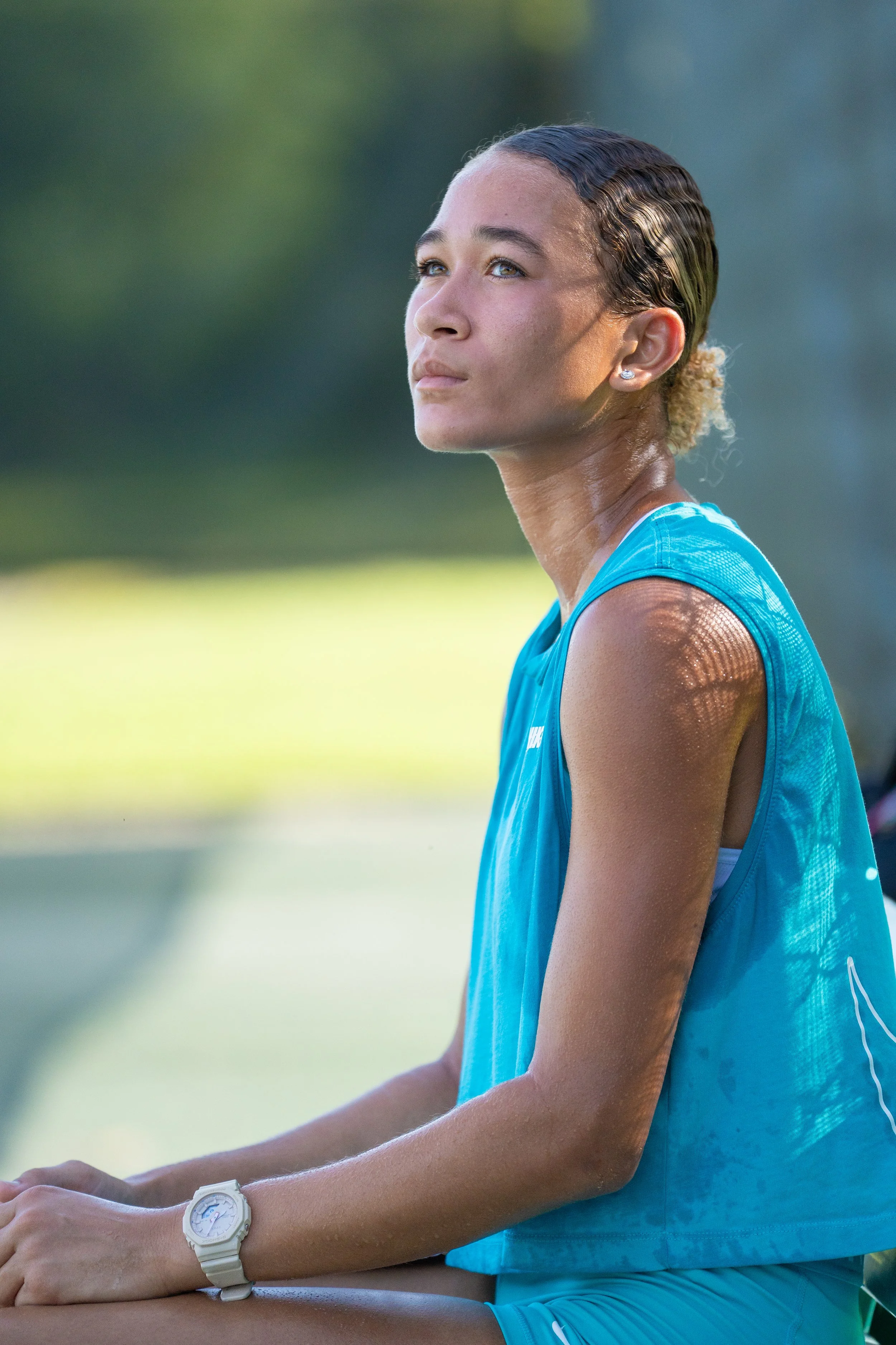 Close-up of a female athlete wearing a blue sports tank top and a white wristwatch, sitting outdoors with a blurred natural background.