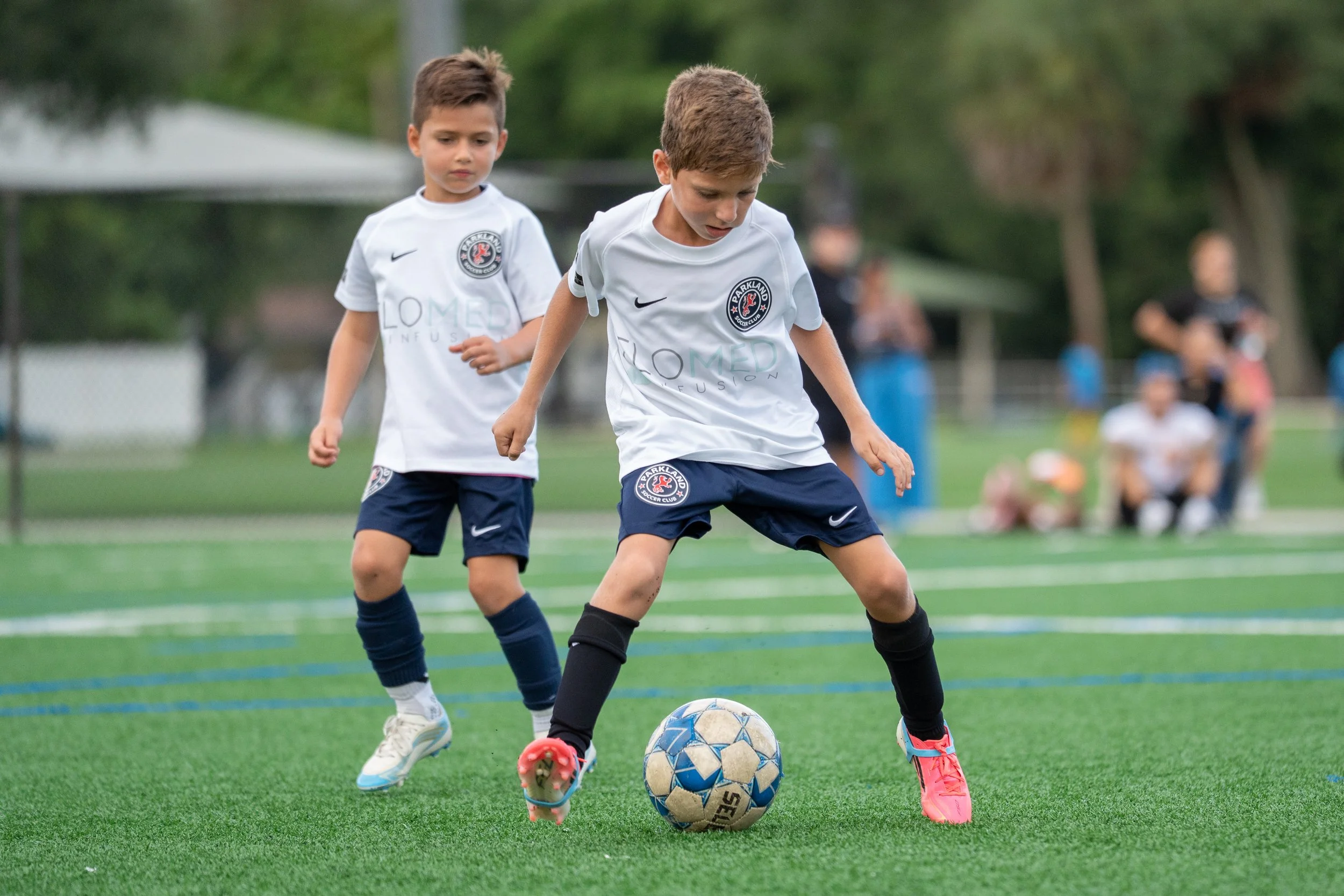 Two young boys wearing white soccer jerseys and blue shorts playing soccer on a field. The boy in front is controlling a blue and white ball with his foot, while the boy behind is watching. In the background, a few people are watching and trees are v