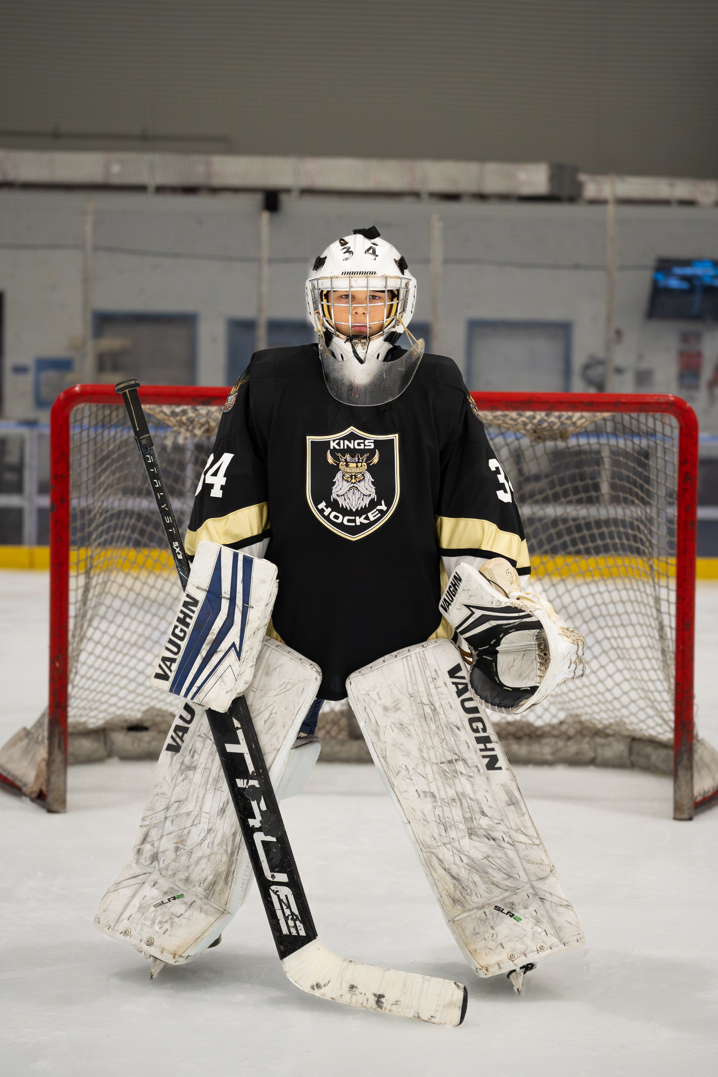 A young hockey goalie in black and gold team uniform standing in front of the goal on an ice rink, wearing a helmet, pads, and holding a hockey stick.