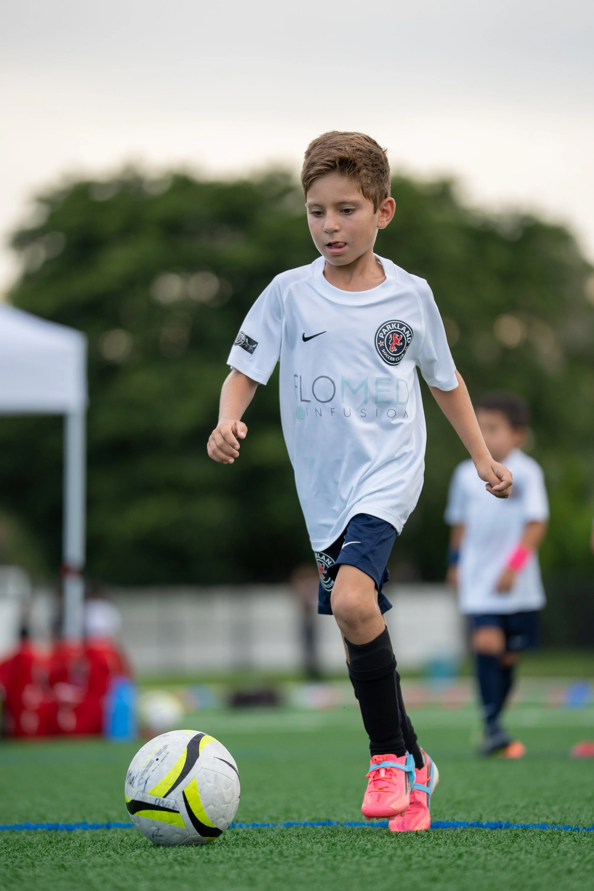 A young boy playing soccer on a field, wearing a white uniform with a team logo, pink shoes, and black socks. He is about to kick a soccer ball, with other children and soccer equipment visible in the background.