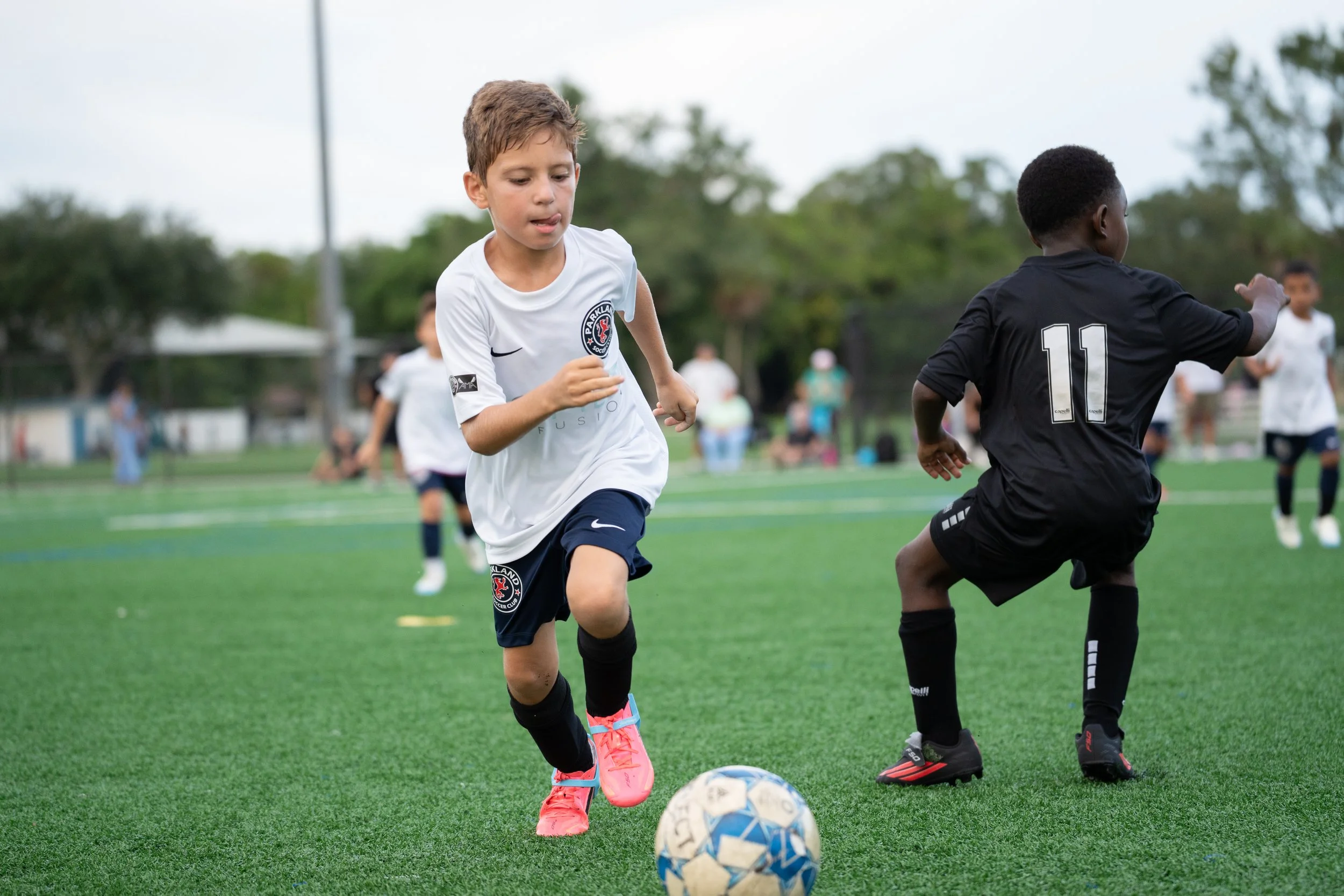Two young boys playing soccer on a field, with one in a white jersey and the other in a black jersey. The boy in white is about to kick a soccer ball, while the boy in black is running nearby.