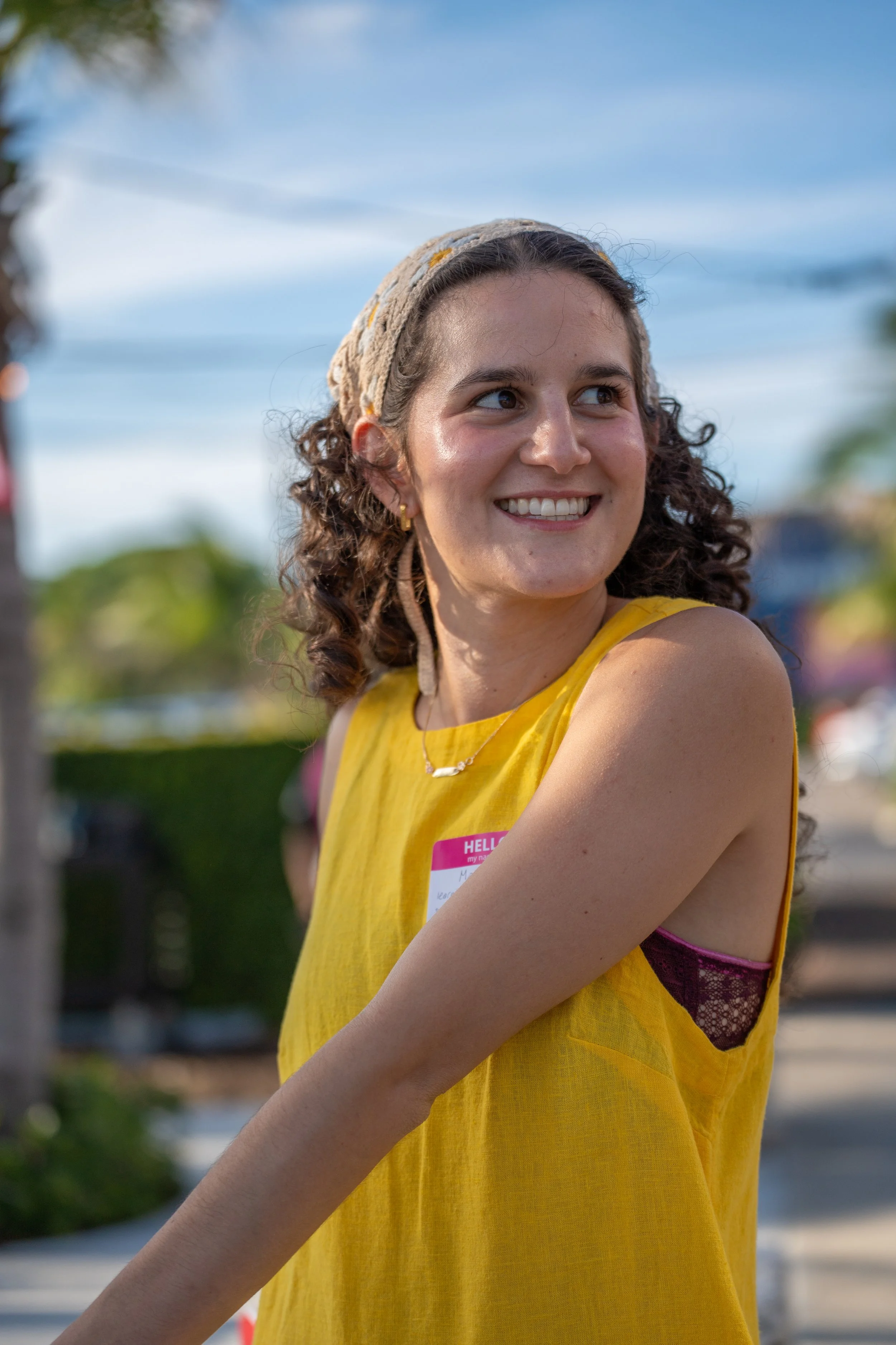 A woman with curly brown hair wearing a yellow sleeveless top and a beige head wrap smiling outdoors.