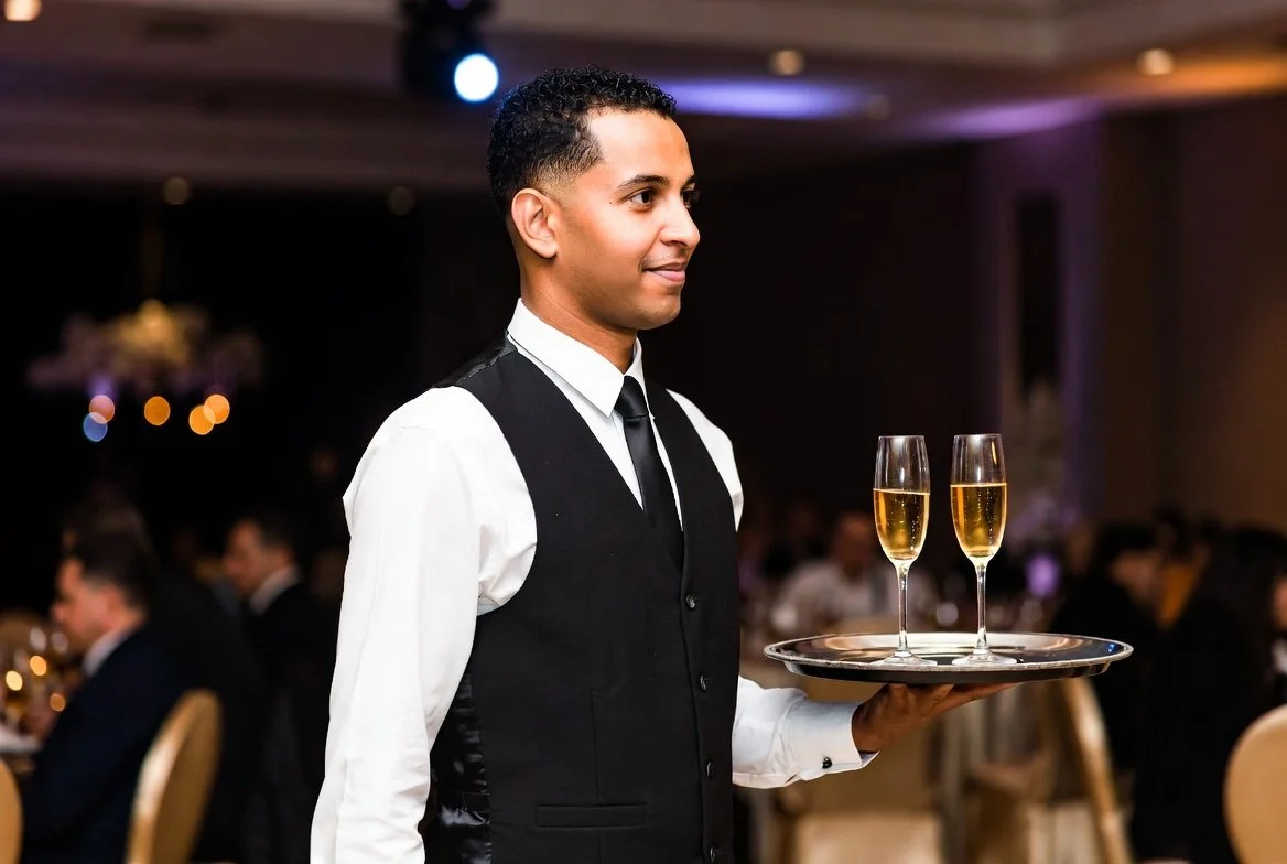 A male waiter in a black vest, white shirt, and black tie carrying a tray with two glasses of champagne at a formal event.