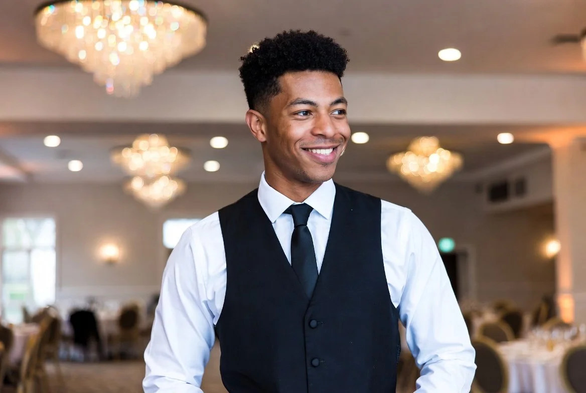 Smiling young man in formal attire at a banquet hall with chandeliers and tables.